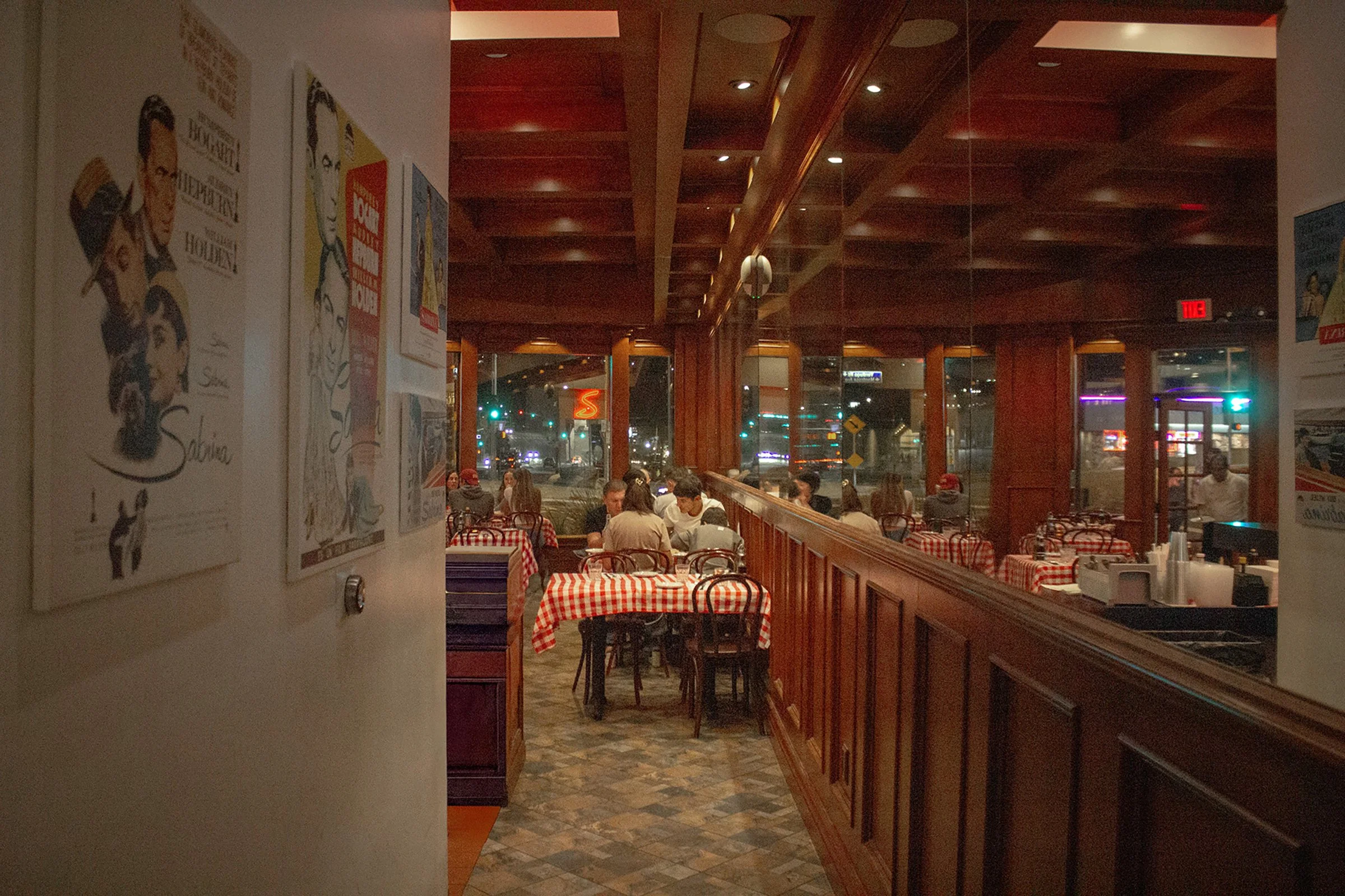 Interior of a cozy restaurant with red and white checkered tablecloths, wooden wall paneling, and large windows showing a city street at night.