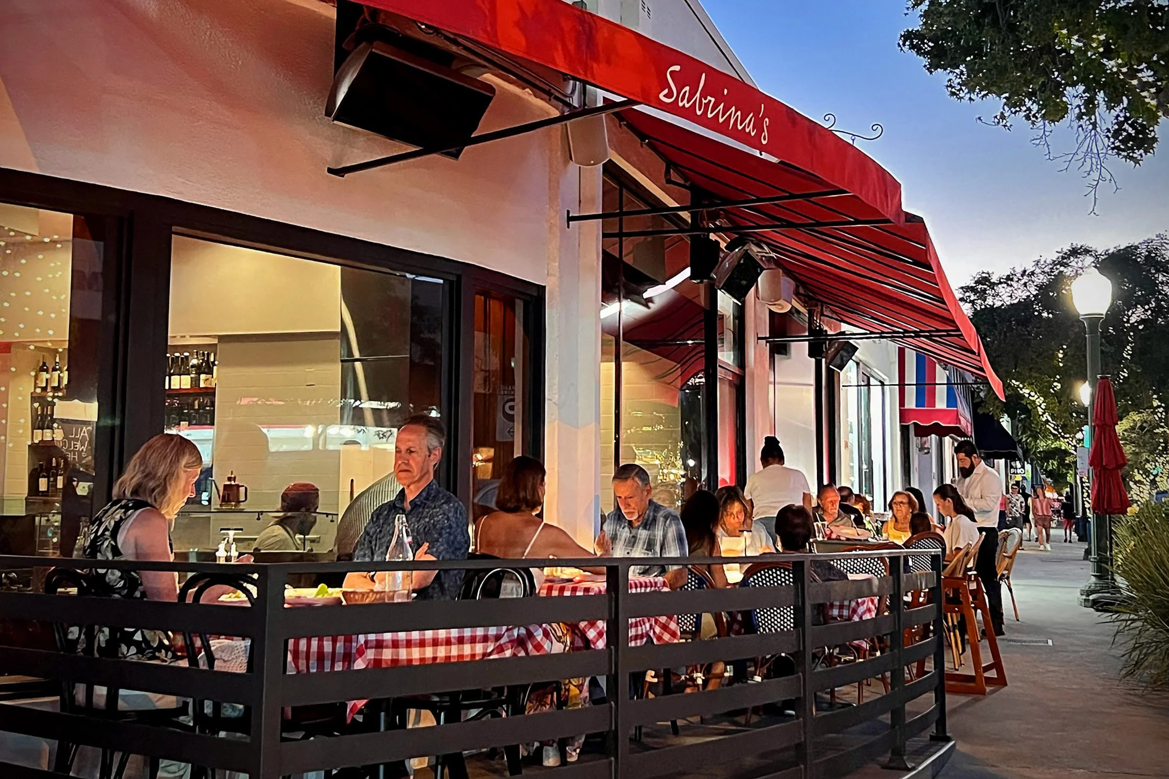 People dining outdoors at Salina's restaurant with red awnings, checkered tablecloths, and string lights on a city sidewalk during evening.