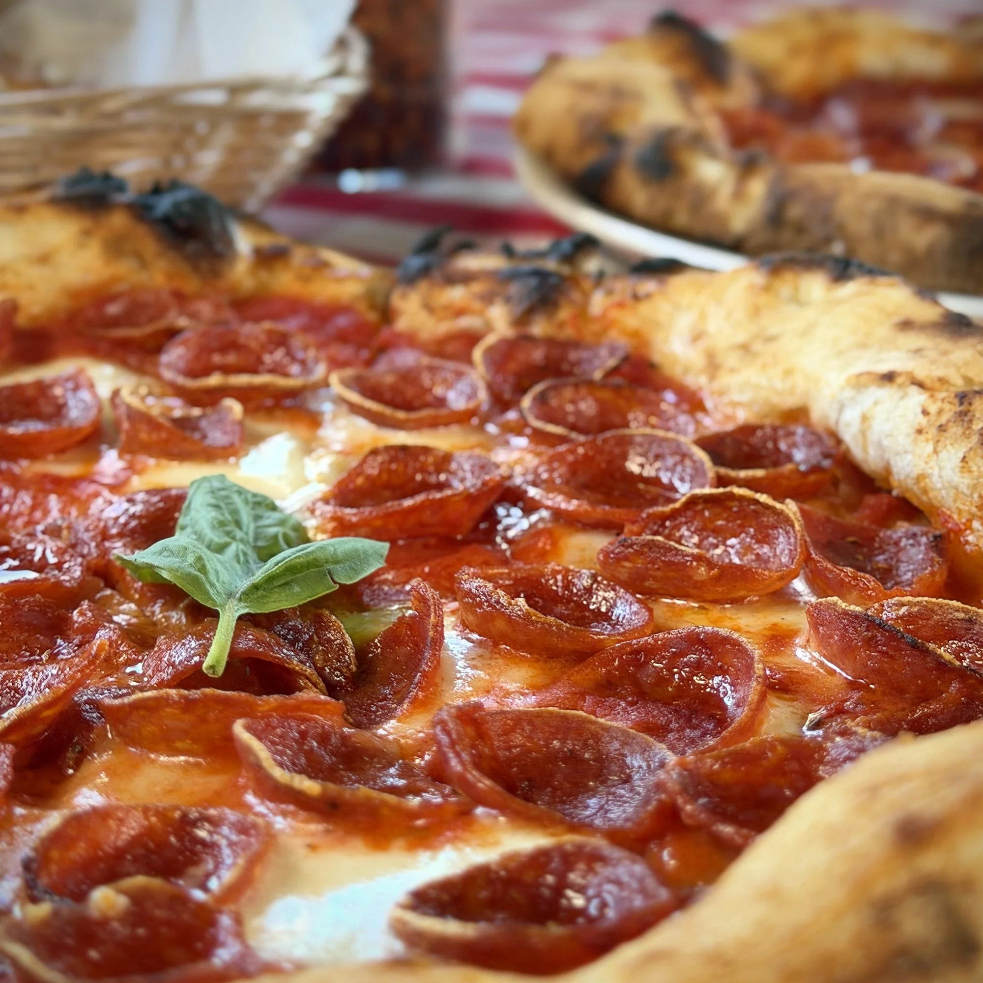 Close-up of a pepperoni pizza with a basil leaf garnish on top, with another pizza in the background