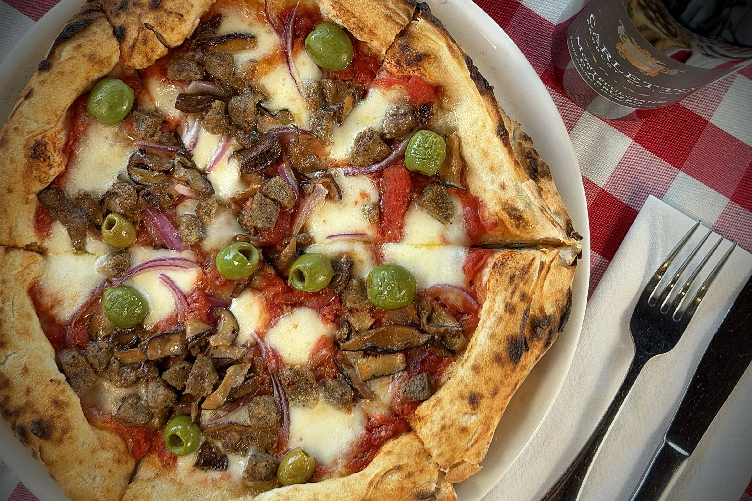 A pizza with mushrooms, green olives, red onions, and melted cheese on a wooden table with a knife, fork, napkin, and a glass of dark soda.
