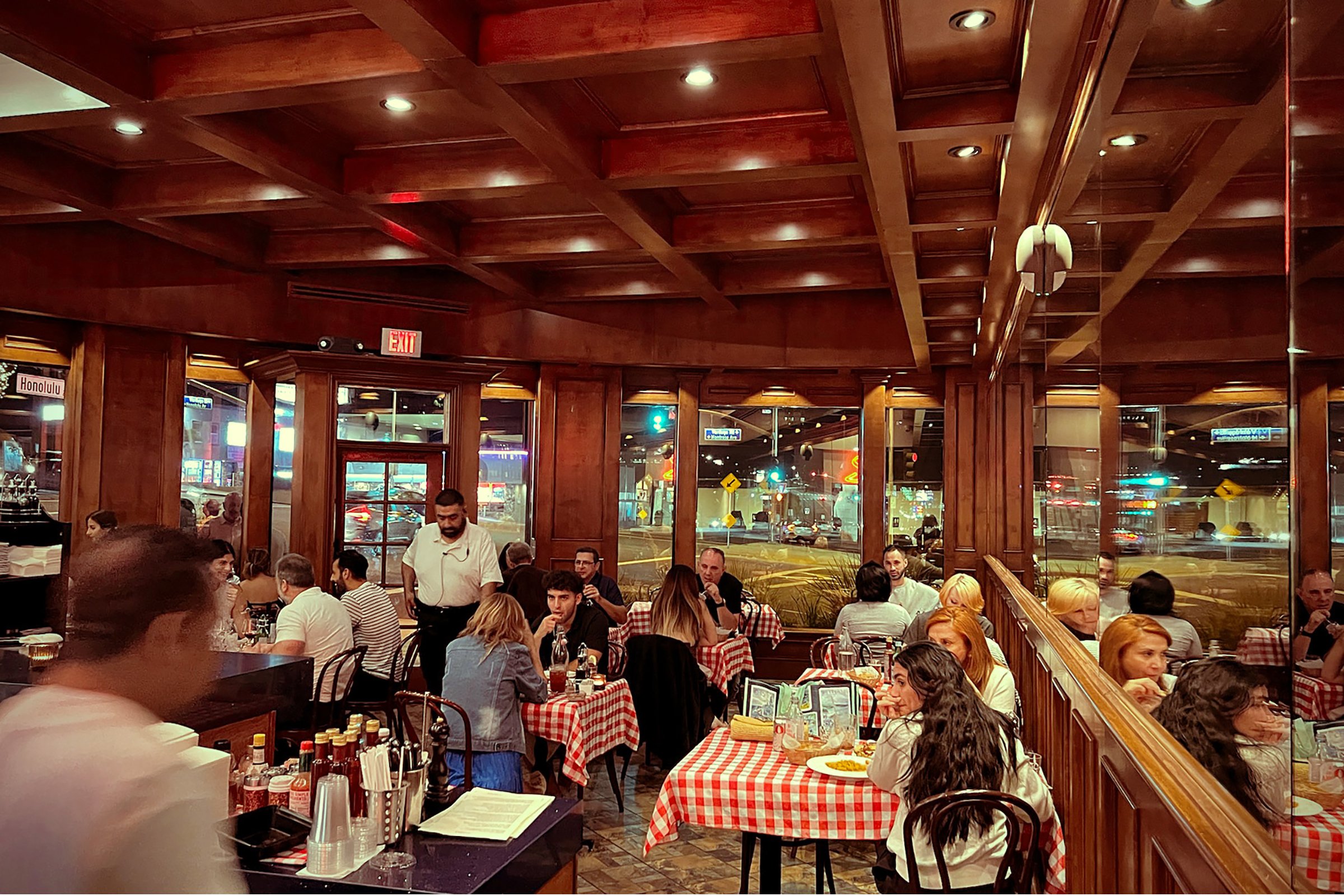 Inside a busy restaurant with wooden decor and checkered tablecloths, several tables are occupied by diners, with a server walking through the room and a window showing a street scene at night.