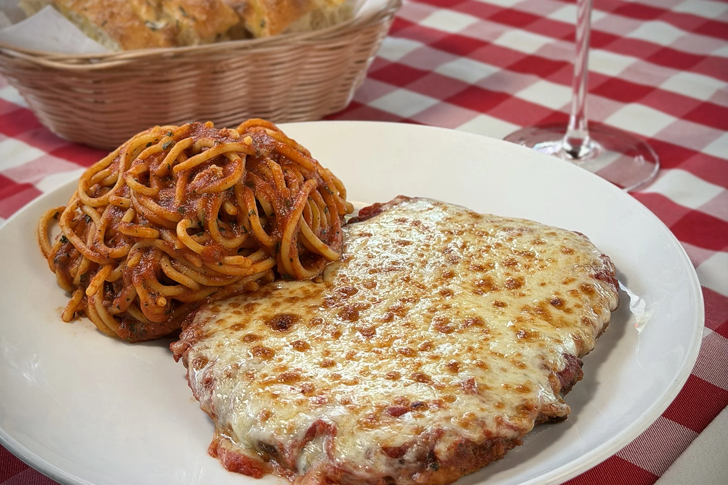 A plate of Italian baked chicken parmesan with spaghetti and marinara sauce, served with a glass of water and a basket of bread in the background.