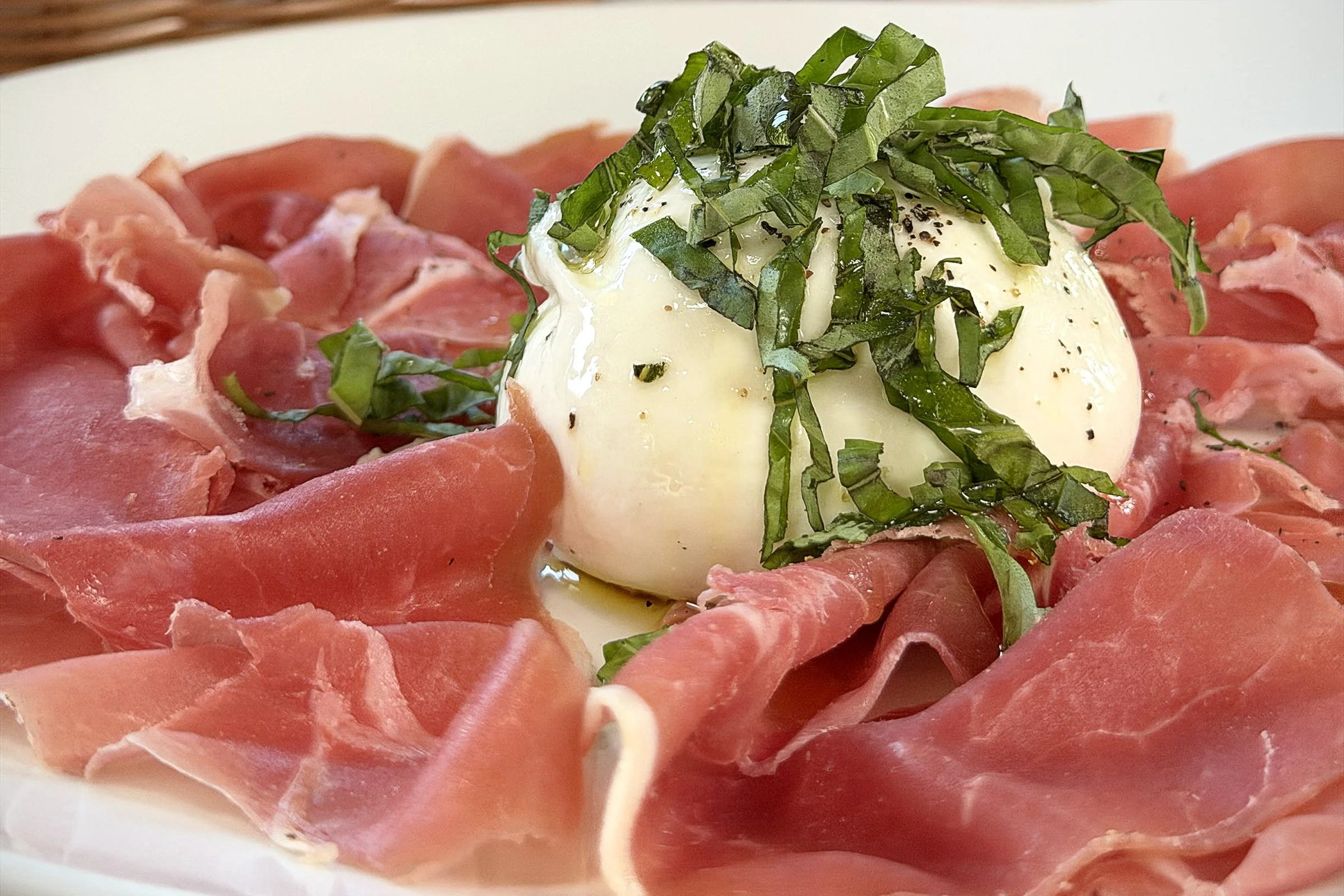 A dish featuring thinly sliced prosciutto, a soft-poached egg topped with chopped basil and black pepper, on a white plate.