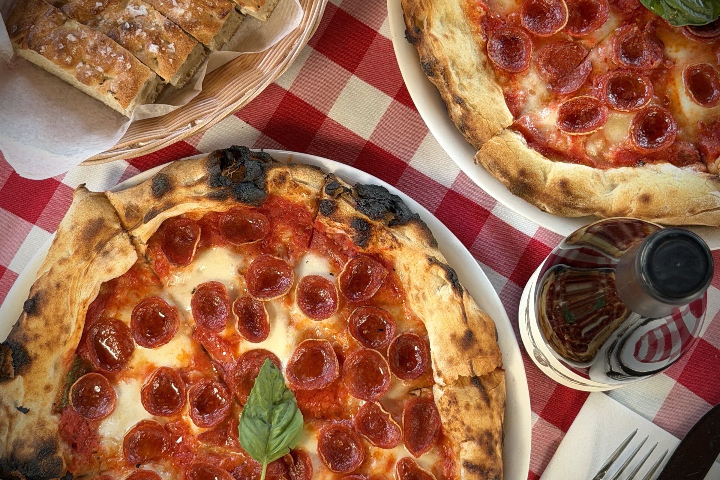Two pepperoni pizzas on a checkered tablecloth with a basket of bread and a bottle of soda.