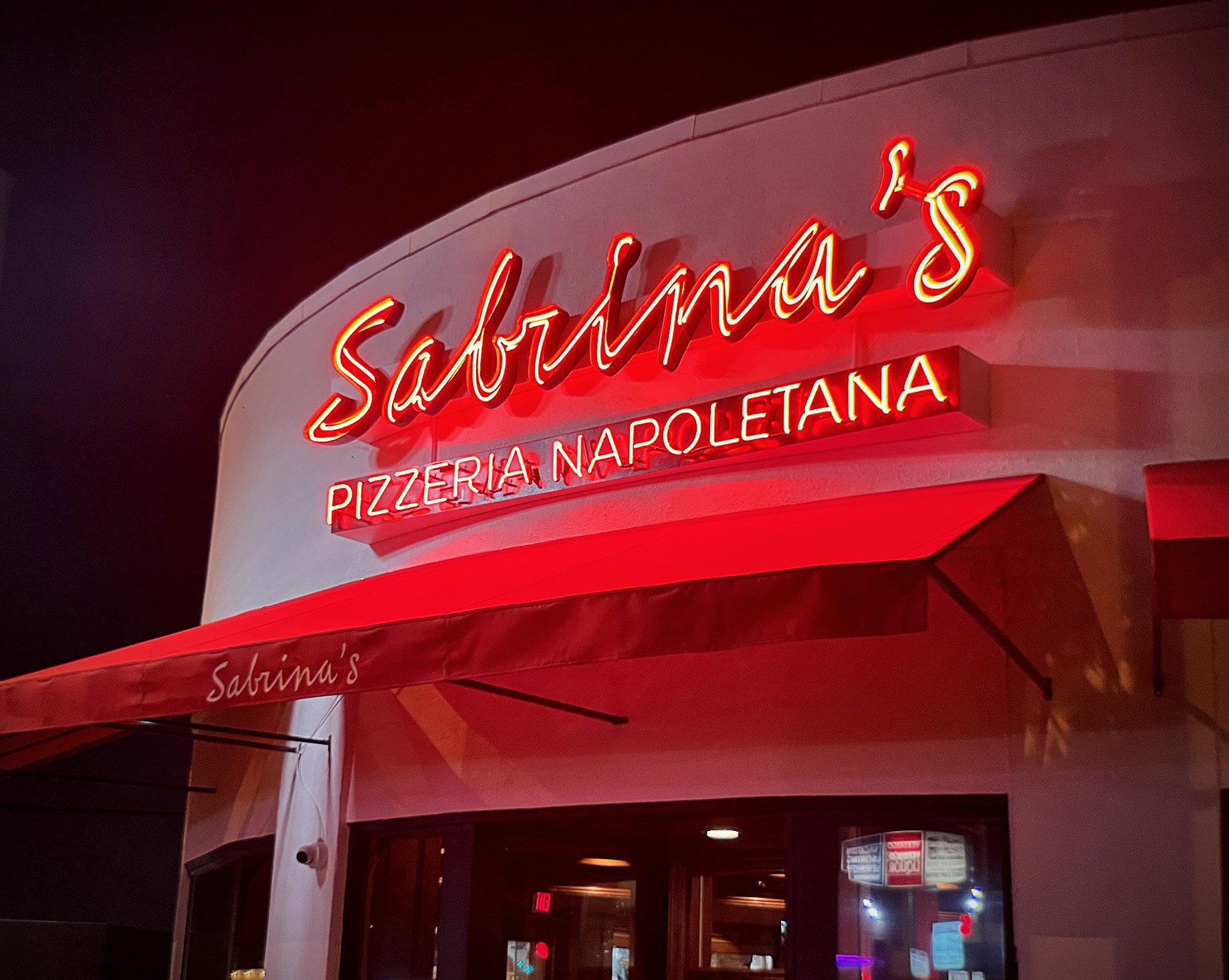 Neon sign outside Sardina's Pizzeria Napolitana at night, with red lights and an awning.