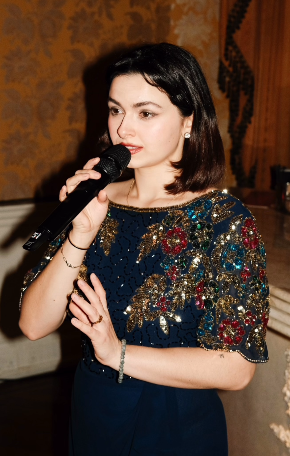 A female jazz singer with dark hair, wearing a vintage dress with gold, red, and green embroidered details, holding a microphone, singing at a private event.