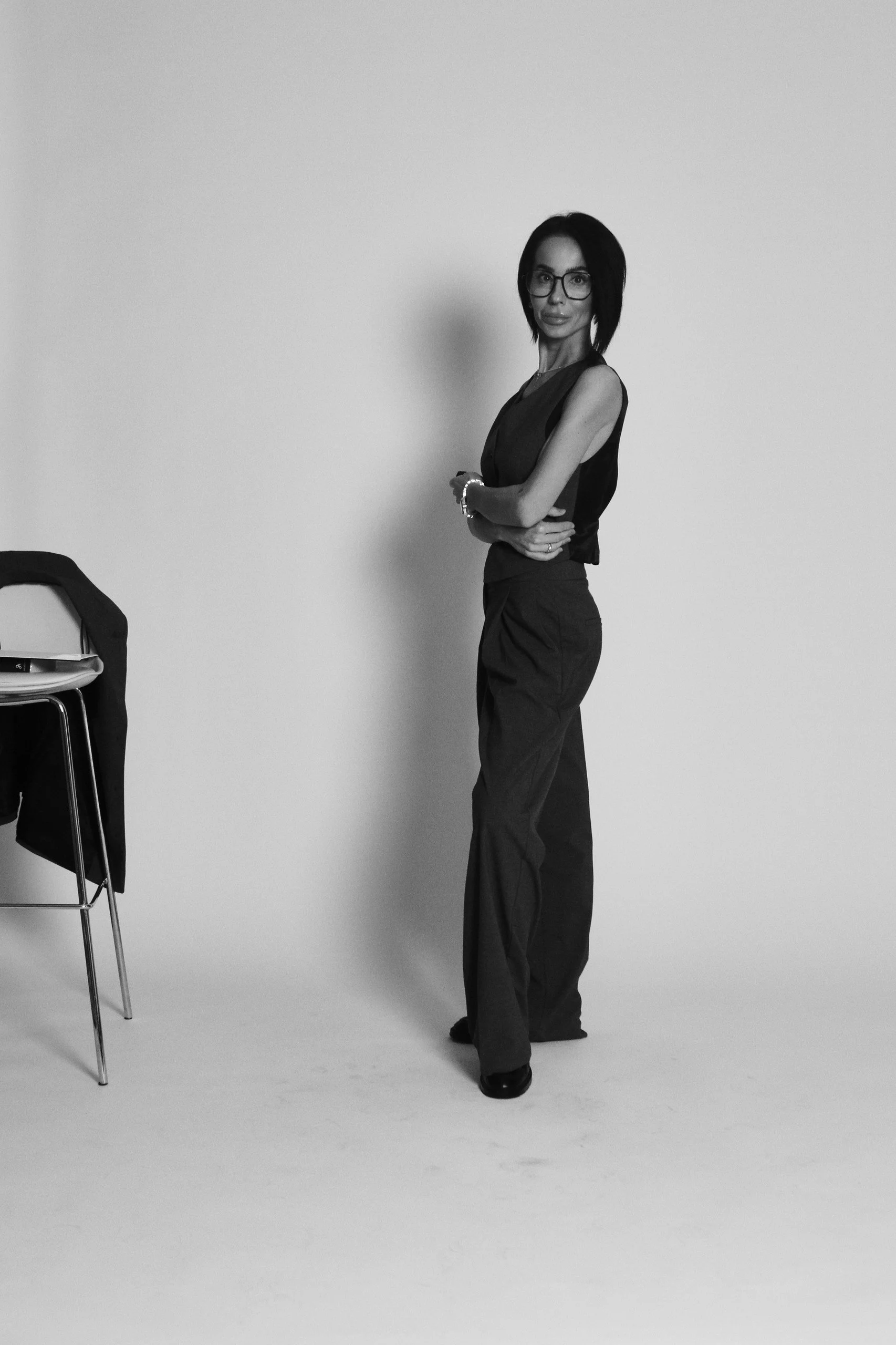 A woman with shoulder-length dark hair, glasses, and light skin, dressed in a sleeveless blouse and wide-leg pants, posing with arms crossed in front of a plain background, with a chair and a jacket on it to her left.