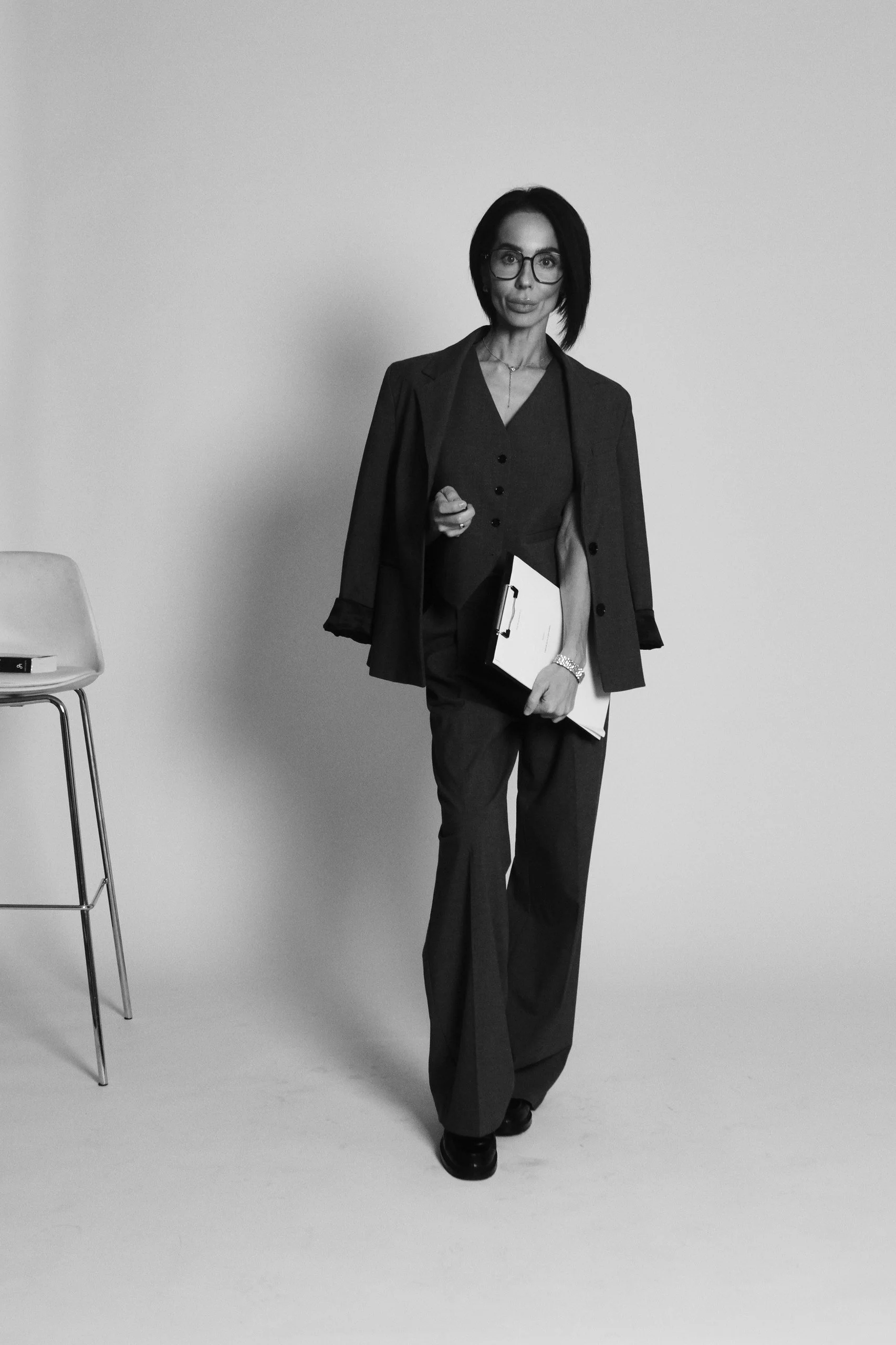 A woman dressed in business attire, wearing glasses and holding a clipboard, standing in front of a plain background.