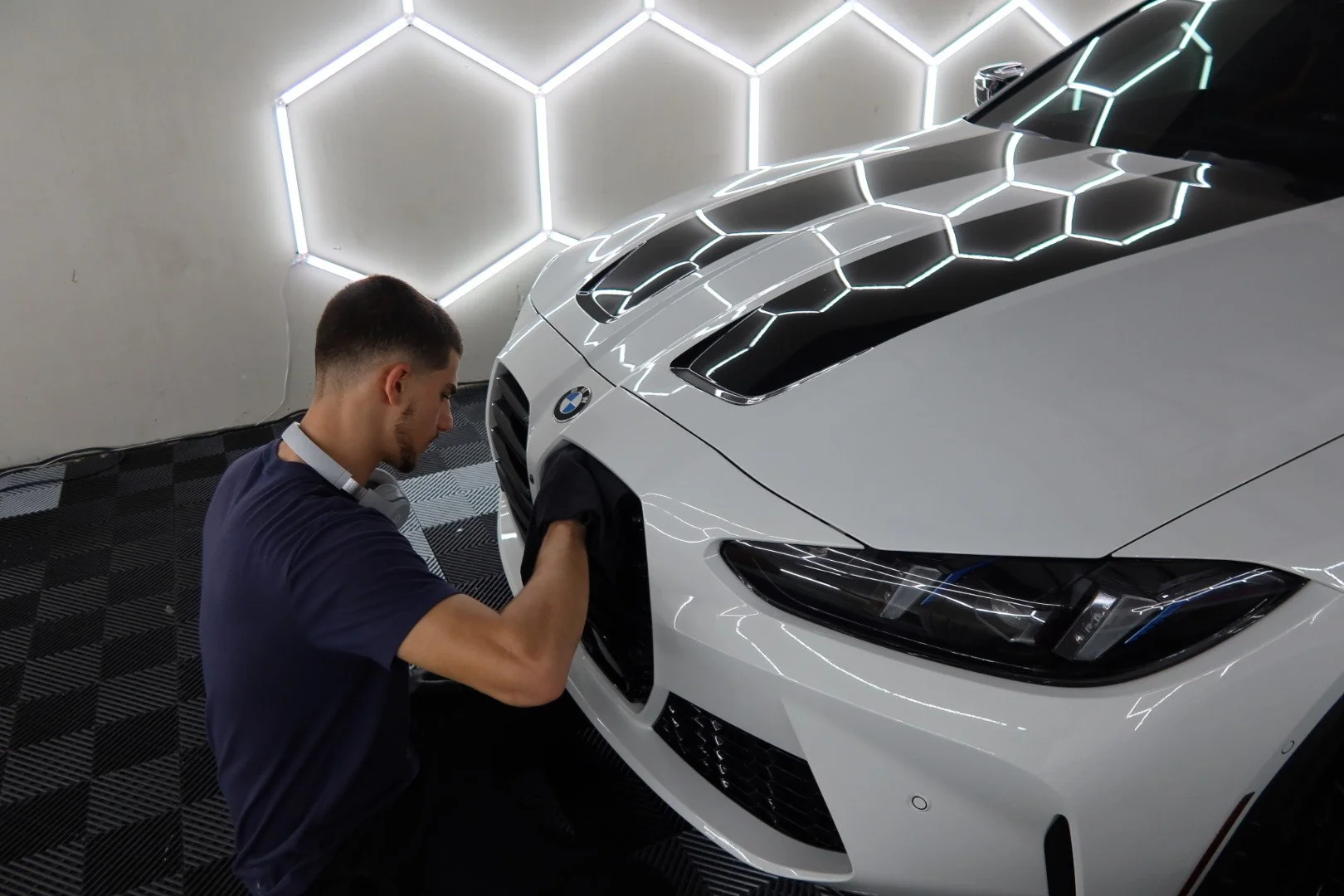 A black high-performance sports car with a large rear wing, being polished by a mechanic wearing gloves in a dark, industrial setting with fluorescent lighting.