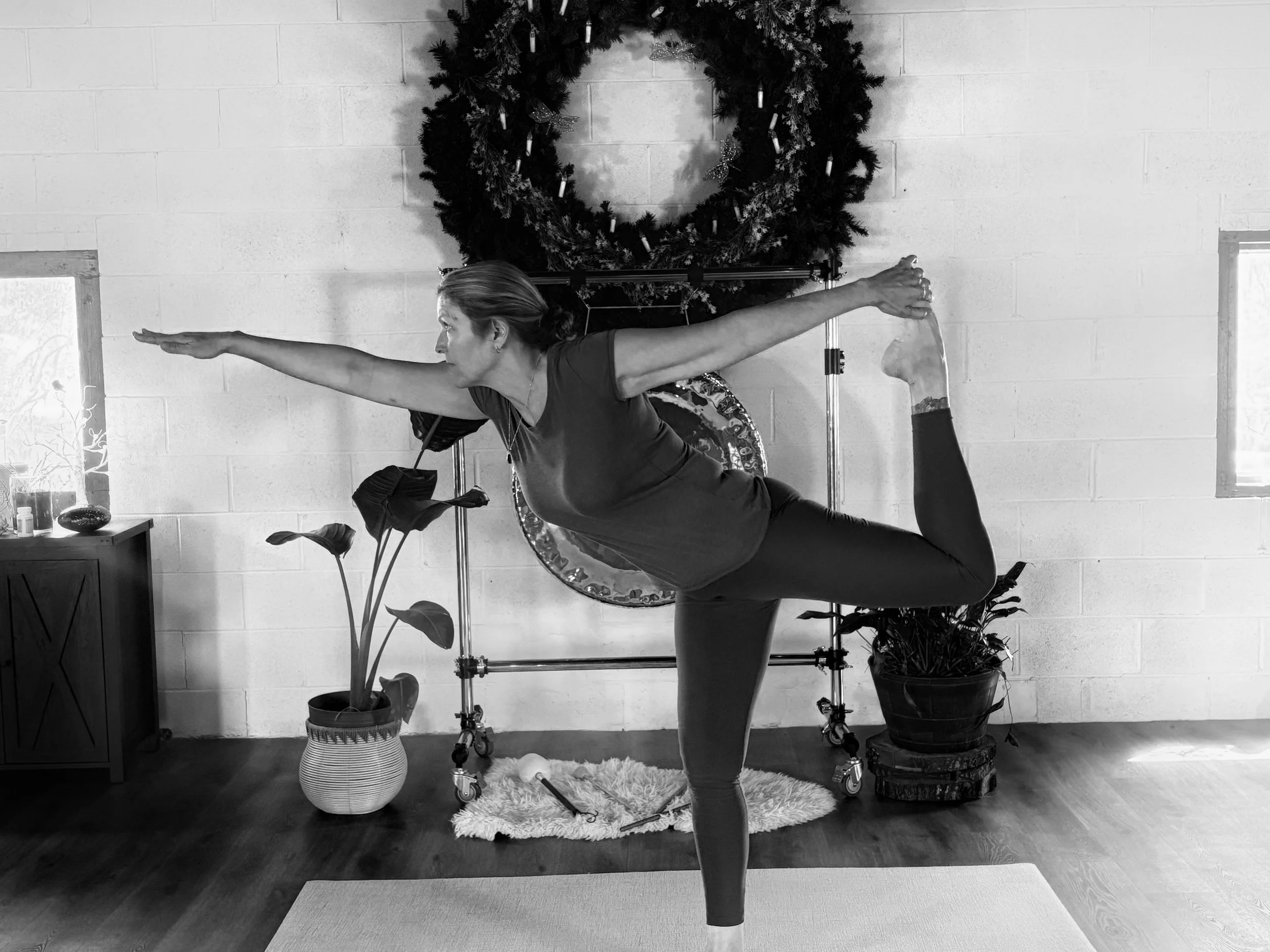Woman practicing yoga indoors, performing a Dancer Pose, with holiday wreath and potted plants in the background.