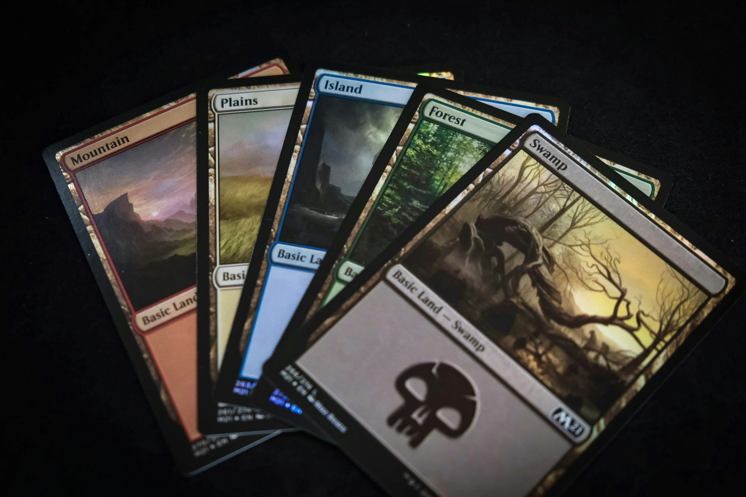 A collection of Magic: The Gathering cards spread out on a black surface, including Mountain, Plains, Island, Forest, and Swamp land cards, with colorful artwork and labels.