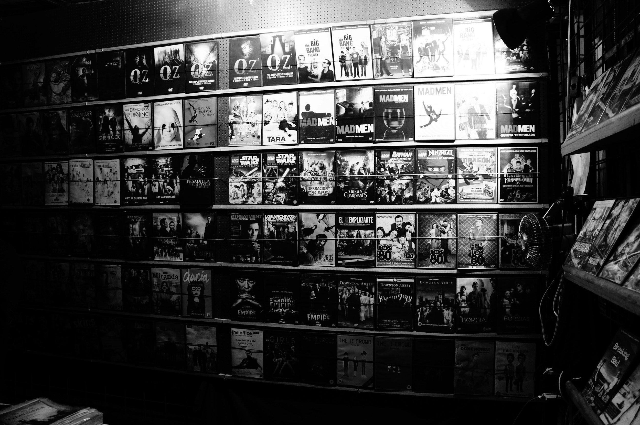 A display of multiple TV show and movie DVD covers arranged on a wall in a store, with several rows of covers visible, including titles like 'Oz', 'Mad Men', 'Star Wars', 'Downton Abbey', and 'The Office'.