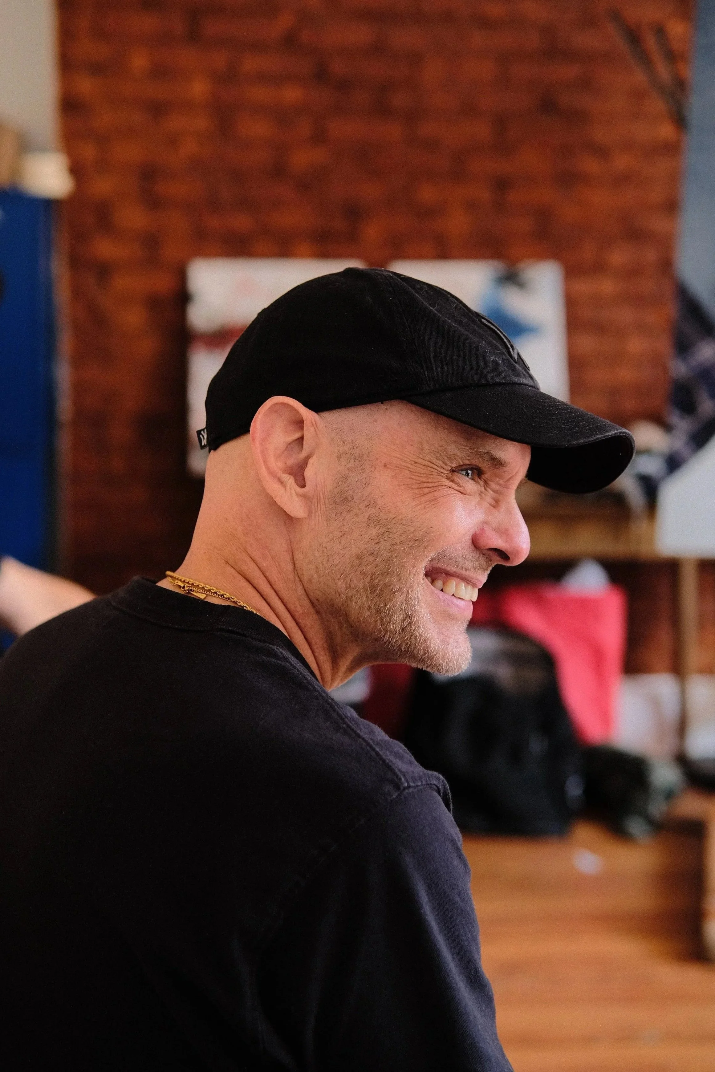 Side profile of a smiling Daniel Flahiff wearing a black cap and black shirt, with a brick wall background and a blurred indoor setting.
