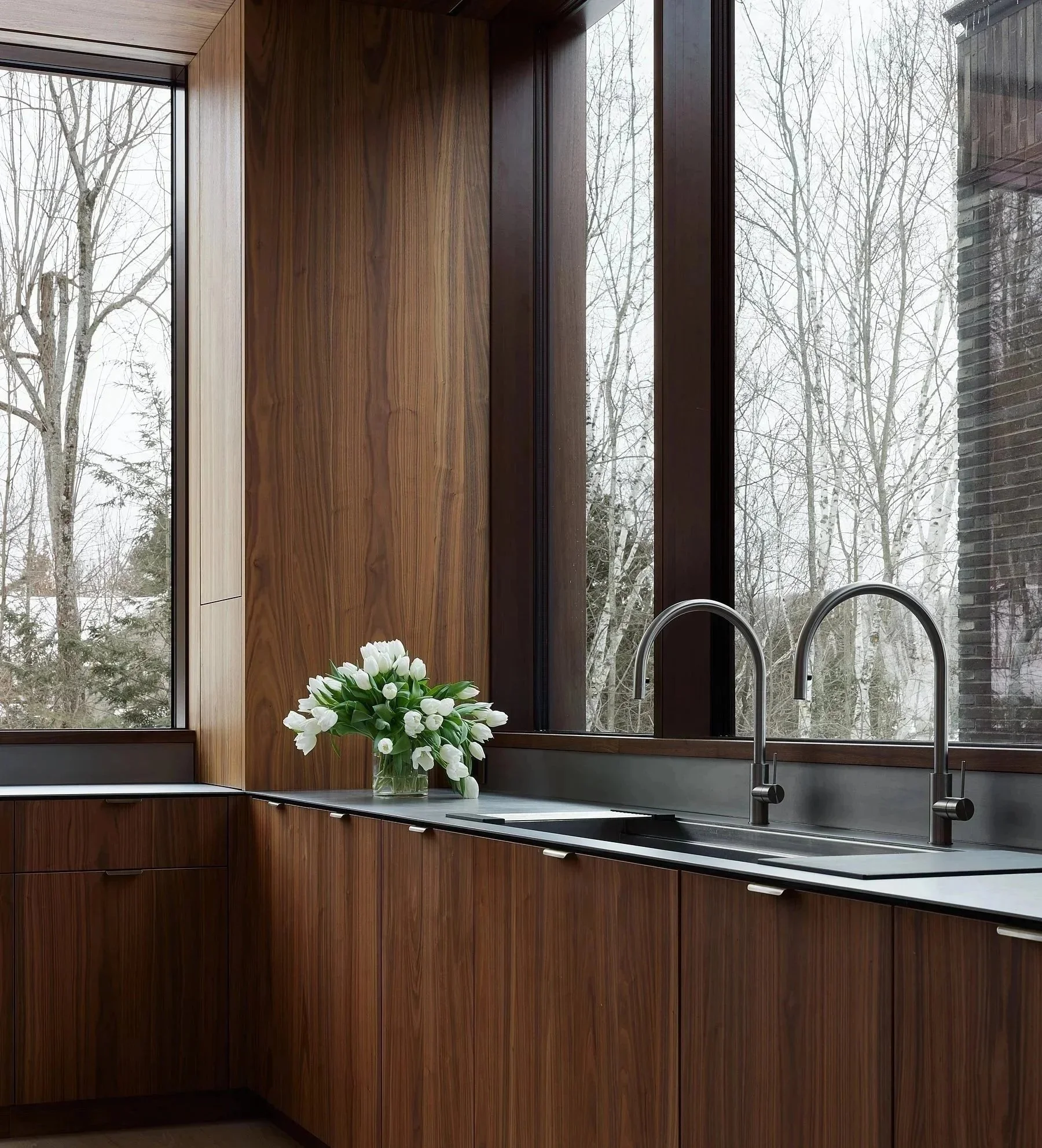 Kitchen with wooden countertop, two faucets, zinc sink, and vase with white tulips, view from a large window with trees outside.