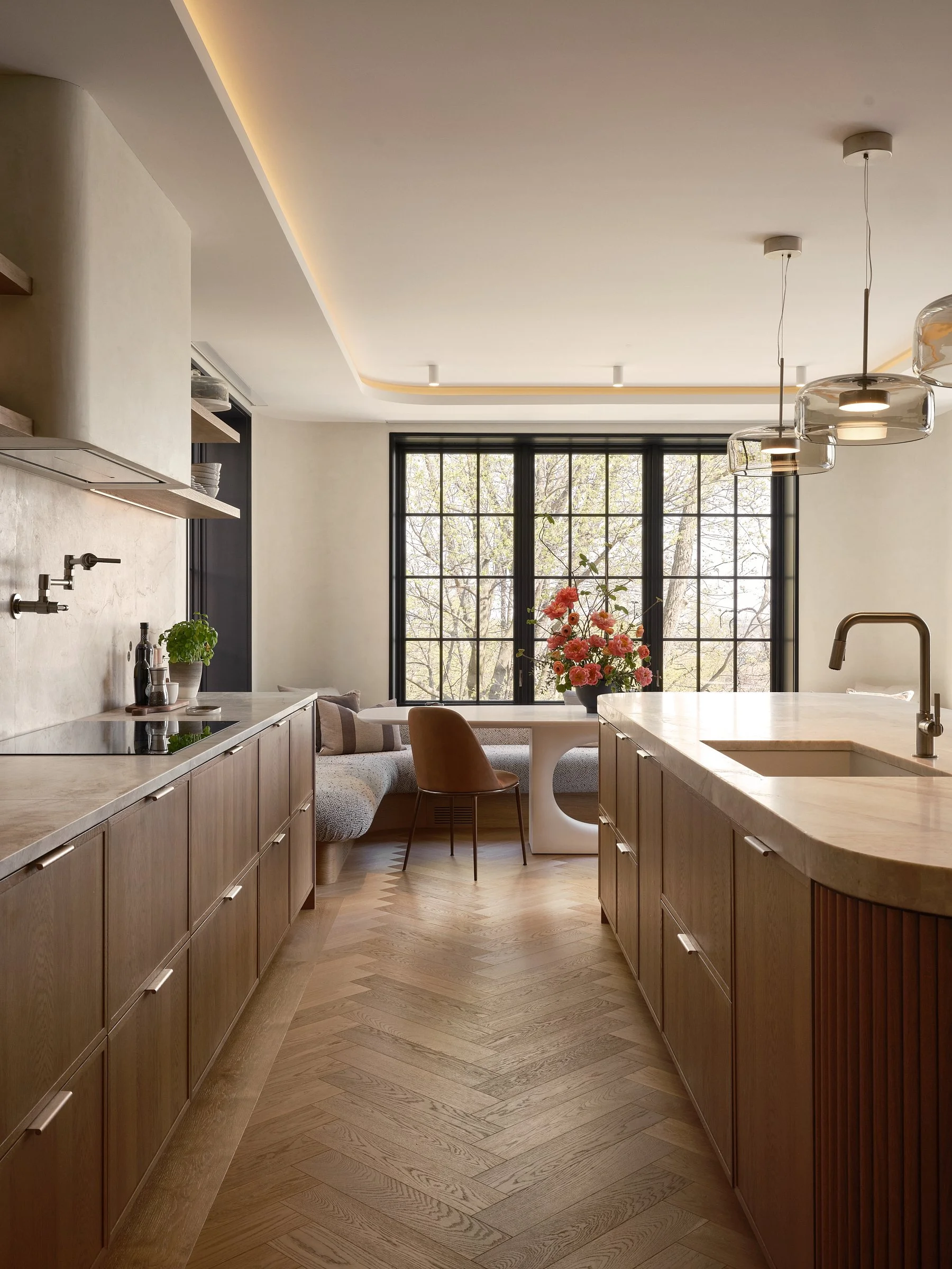 Vue de la cuisine en bois avec îlot en pierre, banquette intégrée et grandes fenêtres donnant sur les arbres.