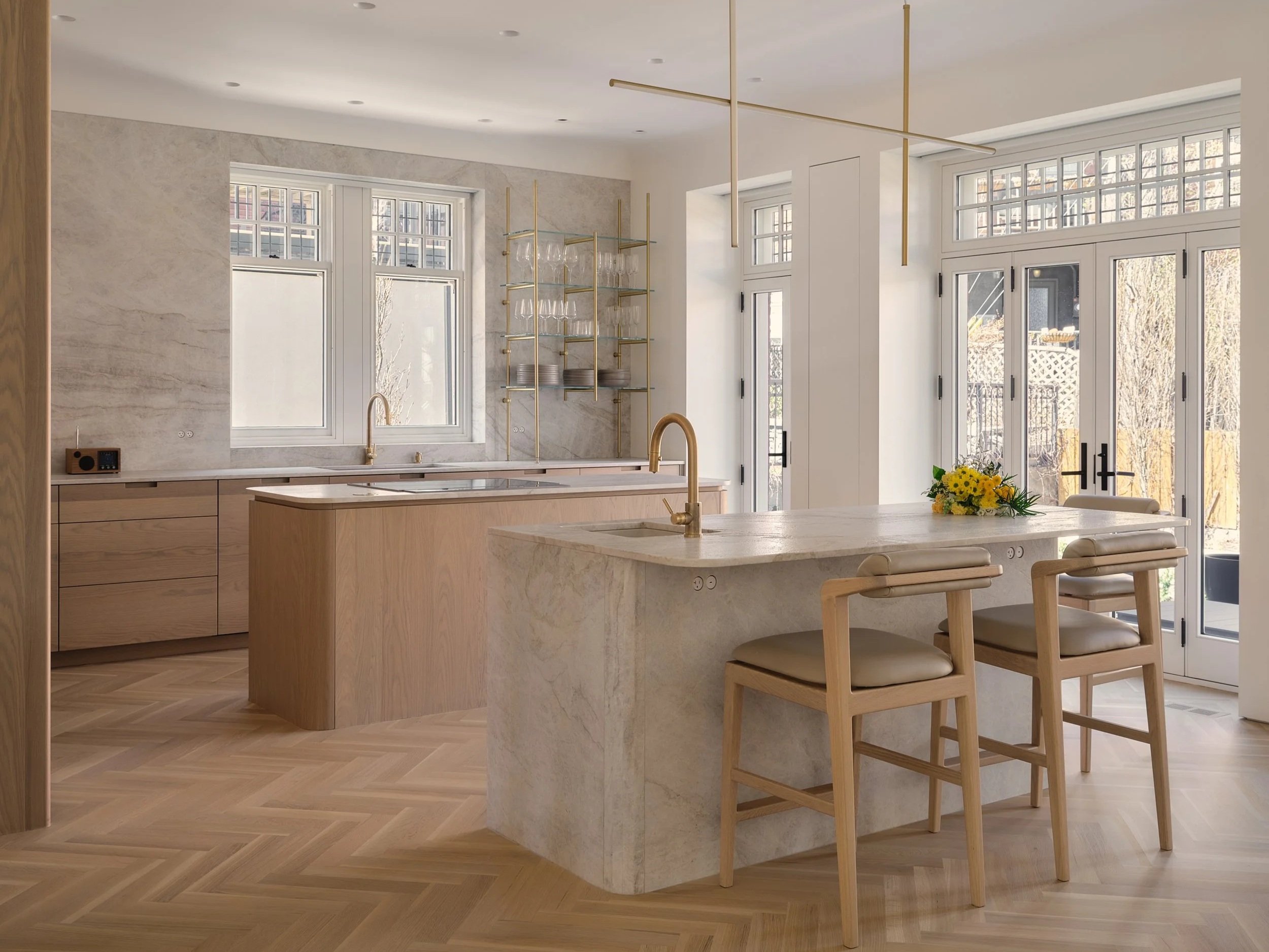 Cuisine moderne avec îlot en marbre, chaises en bois, grandes fenêtres laissant entrer la lumière naturelle, étagère en métal doré avec verres et assiettes, sol en parquet en chevrons.