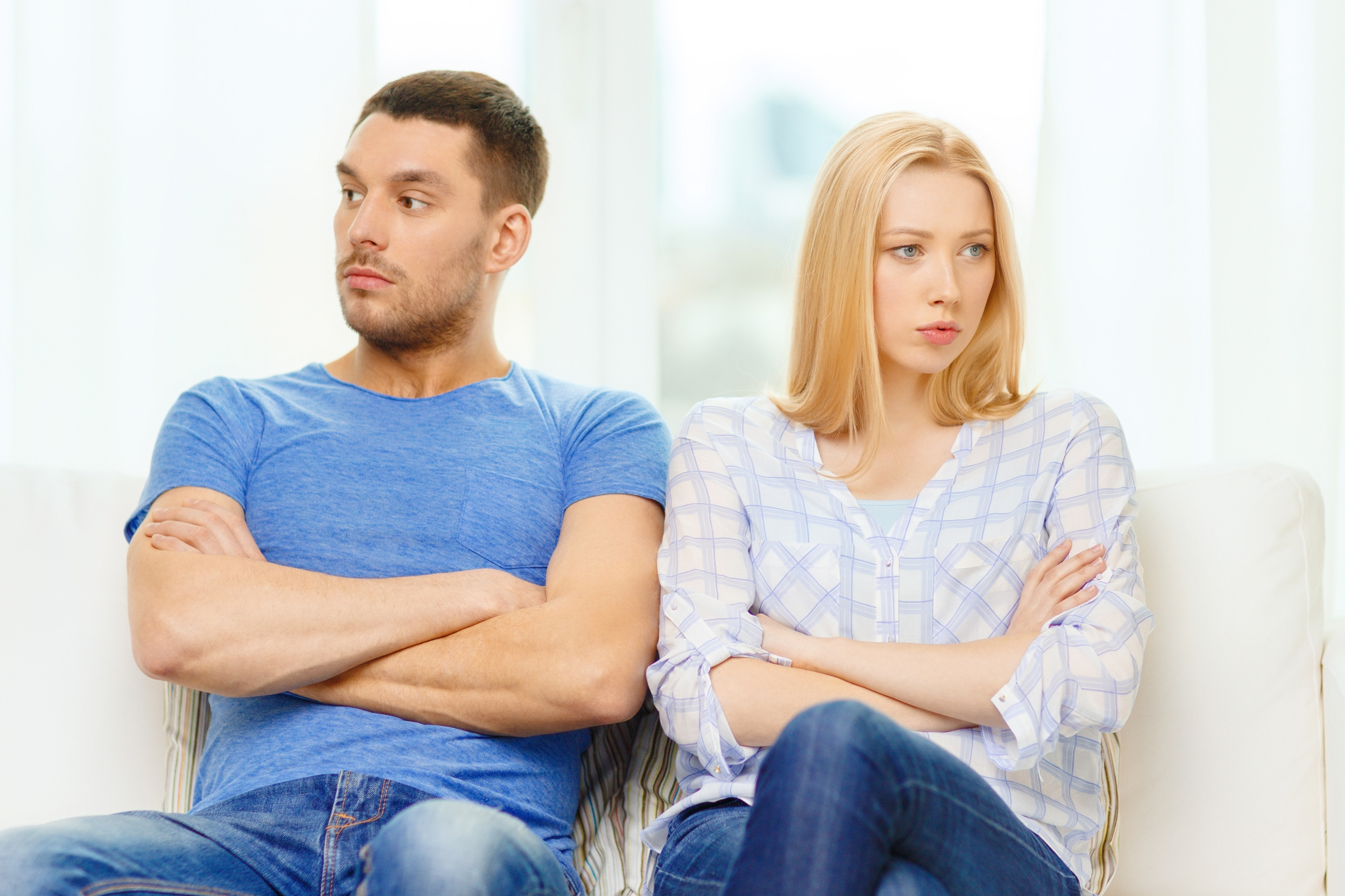 A young man and woman sitting on a white sofa facing away from each other with crossed arms and upset expressions, highlighting relationship issues resolved through couples therapy in North Richland Hills.