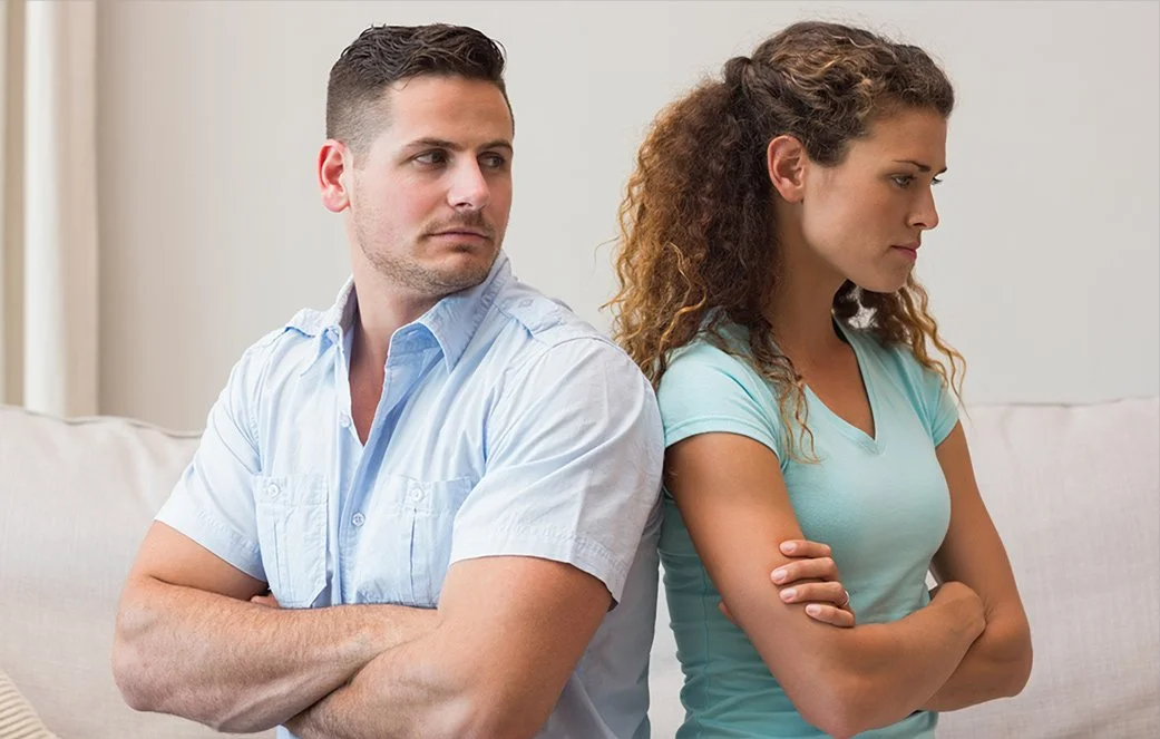 An unhappy man and woman sitting back-to-back on a couch with crossed arms, illustrating the relationship distance and conflict addressed in couples counseling in North Richland Hills.