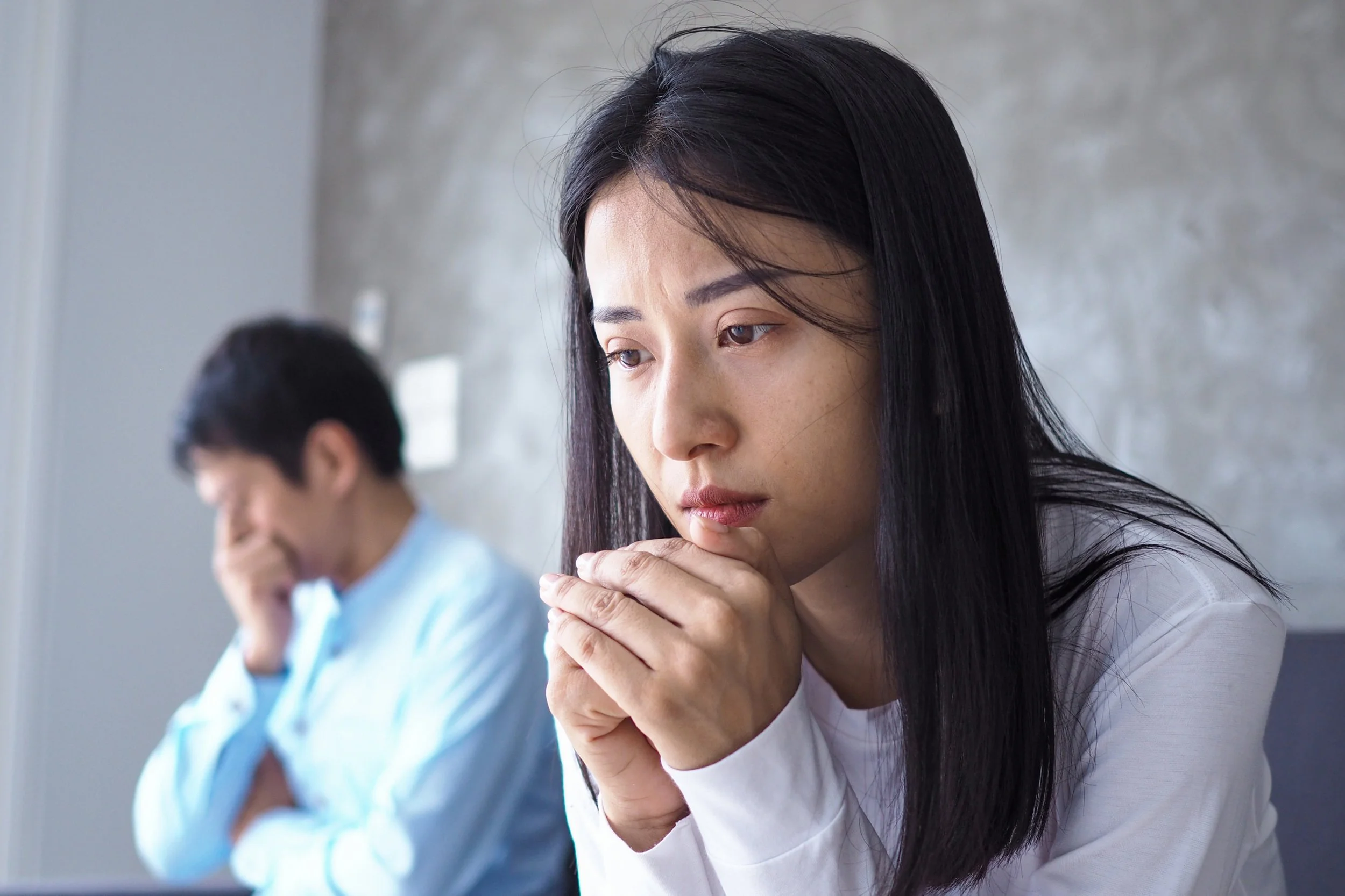 A close-up of a worried Asian woman with clasped hands sitting near a blurred, stressed man on a couch, representing the common marital distress addressed through couples counseling and marriage therapy.