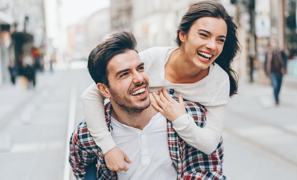 A joyful young man giving a piggyback ride to a laughing woman on a city street, showcasing a happy, connected relationship restored through couples counseling in Colleyville.