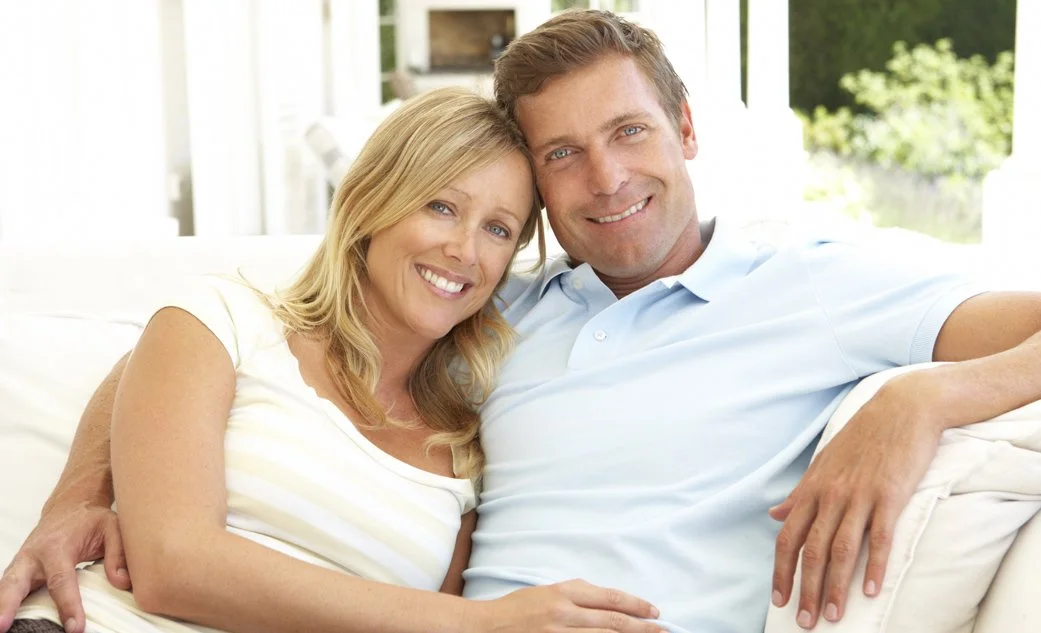 A happy, smiling blonde woman and her husband relaxing together closely on a white outdoor sofa, illustrating a reconnected relationship built through couples counseling in Keller.