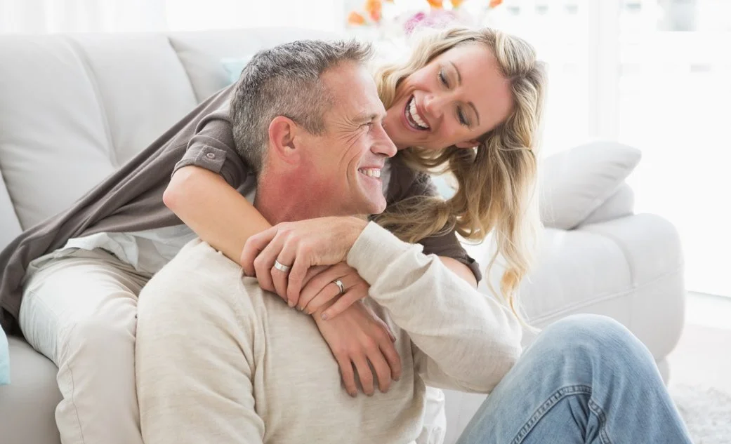 A smiling woman leaning over the back of a sofa to embrace her happy husband from behind, demonstrating the joy and deep connection restored through couples counseling in Southlake.