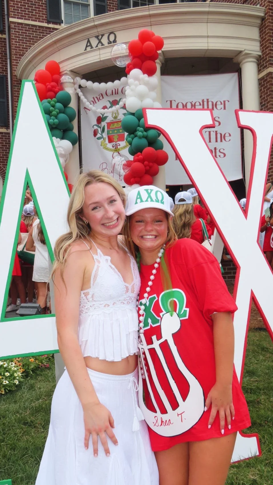 Two women smiling in front of a graduation event backdrop with large Greek letters, balloons, and a banner. One woman in white, the other in red, wearing graduation accessories with Greek letters.