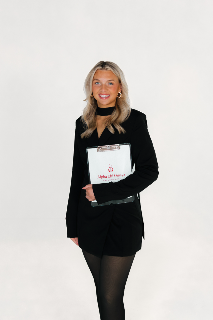 A young woman with blonde hair wearing a sleeveless white top with gold buttons, light blue jeans, and jewelry, sitting on a brick ledge outside near a brick wall and a door with a sign that reads 'Caution Low Clearance'.