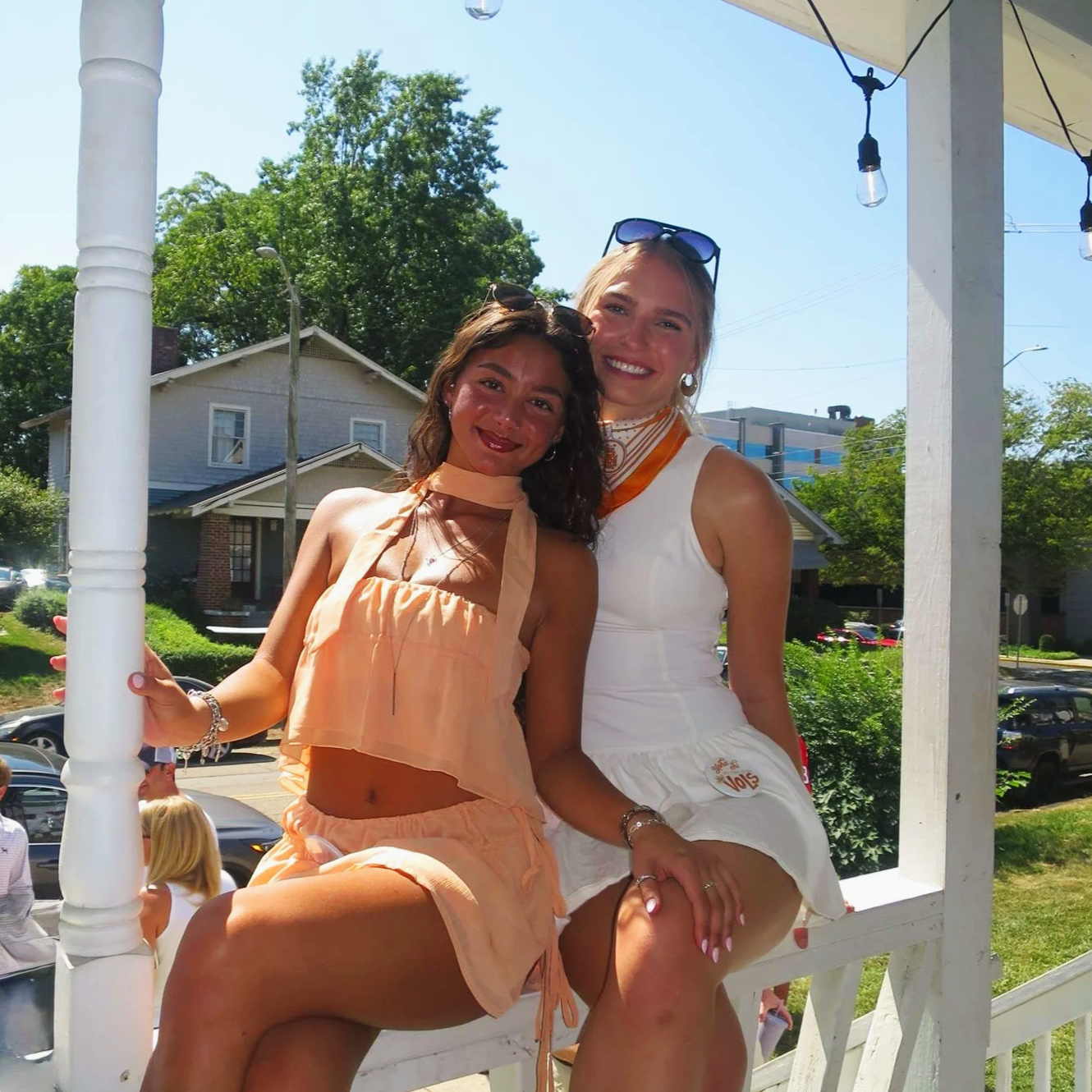 Two women sitting closely together on a white porch railing outdoors, smiling at the camera, with a house, trees, and parked cars in the background on a sunny day.