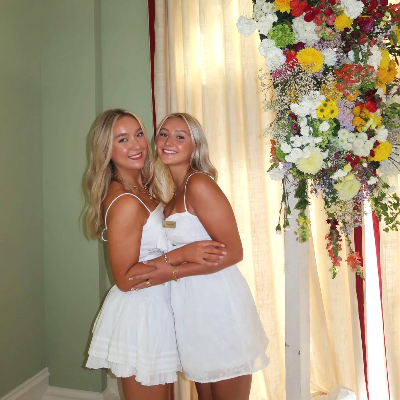 Two young women in white dresses smiling and hugging each other, standing in front of a large colorful floral arrangement and sheer curtains.