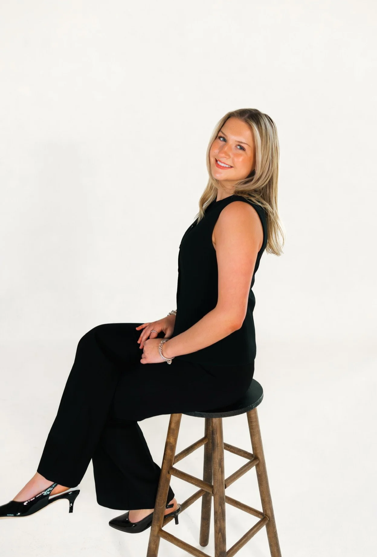 A woman with long blonde hair smiling, wearing a red sleeveless dress, standing in front of a dark striped background.