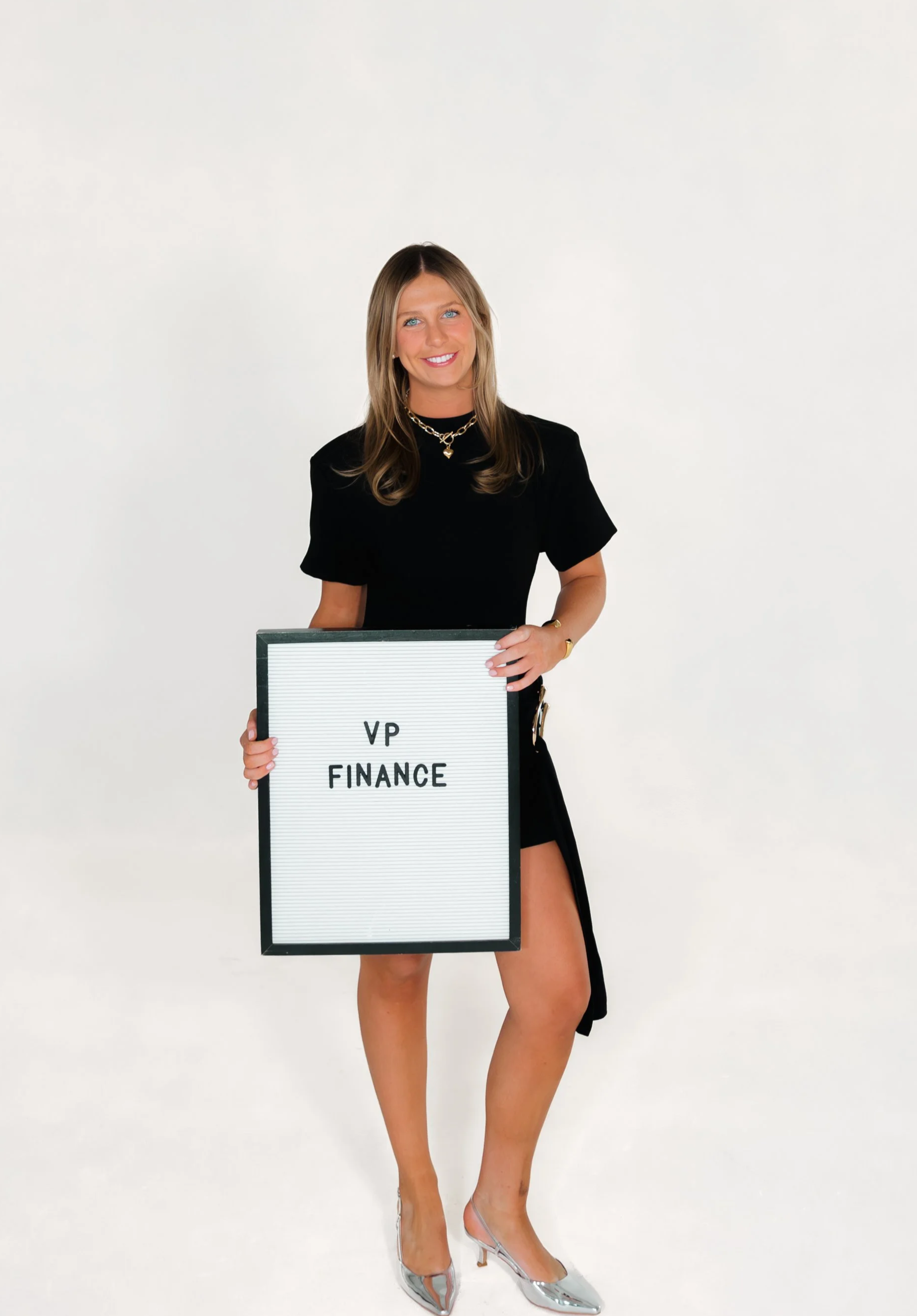 A woman with blonde hair smiling outdoors, wearing a white spaghetti strap top and gold jewelry, with green trees in the background.