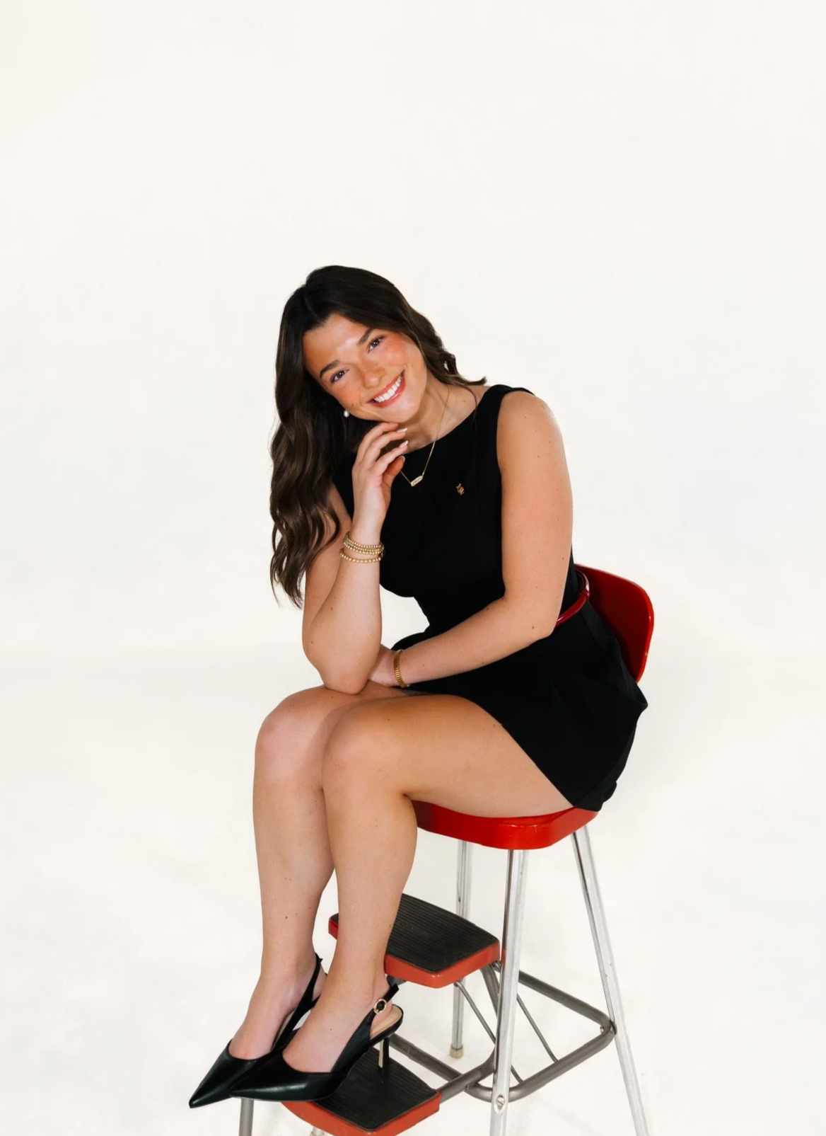 Smiling young woman with long brown hair in loose waves, standing in front of a stone wall.