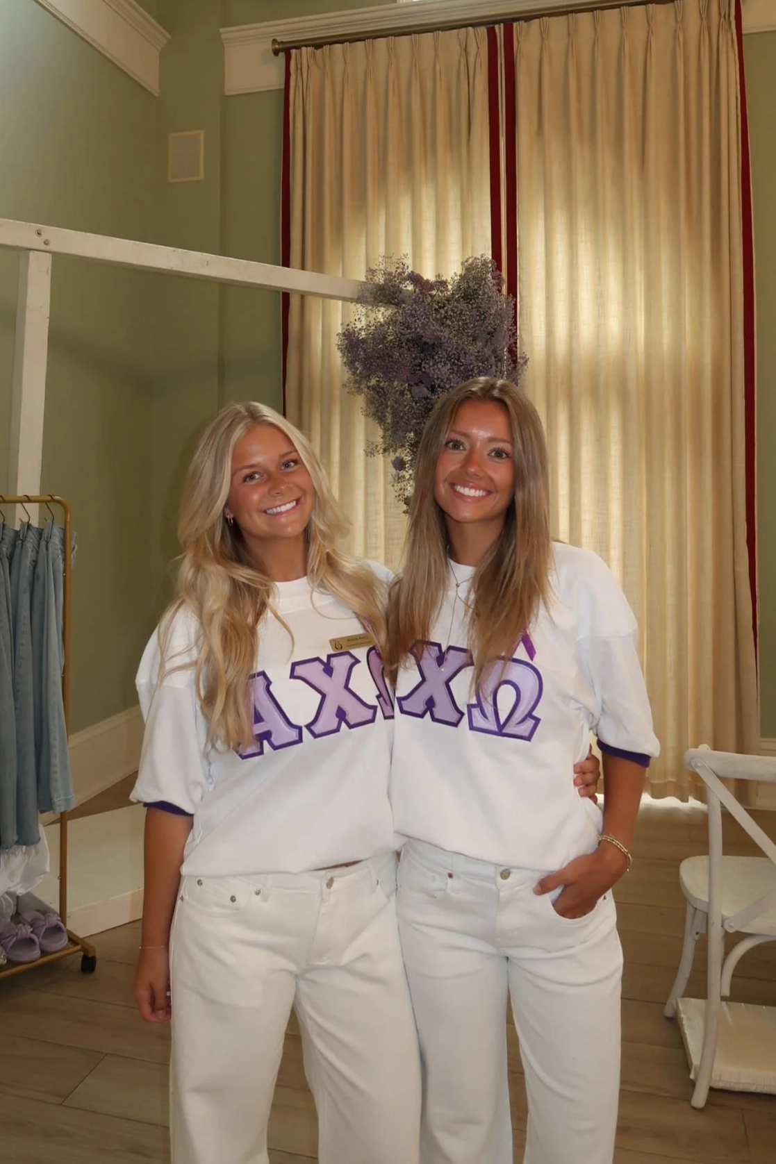 Two young women wearing white Sigma Sigma Sigma sorority t-shirts, smiling, standing in a room with beige curtains and wooden floors.