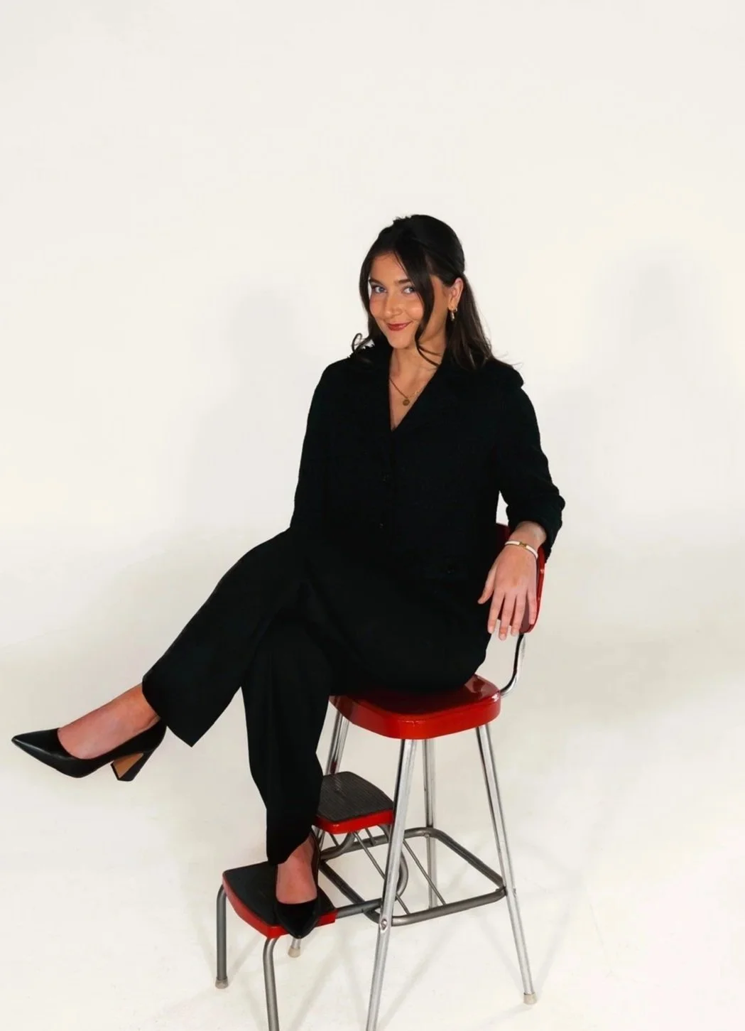 A young woman with long, wavy brown hair smiling and tilting her head in a grassy field with trees and hills in the background, wearing a white sleeveless dress.