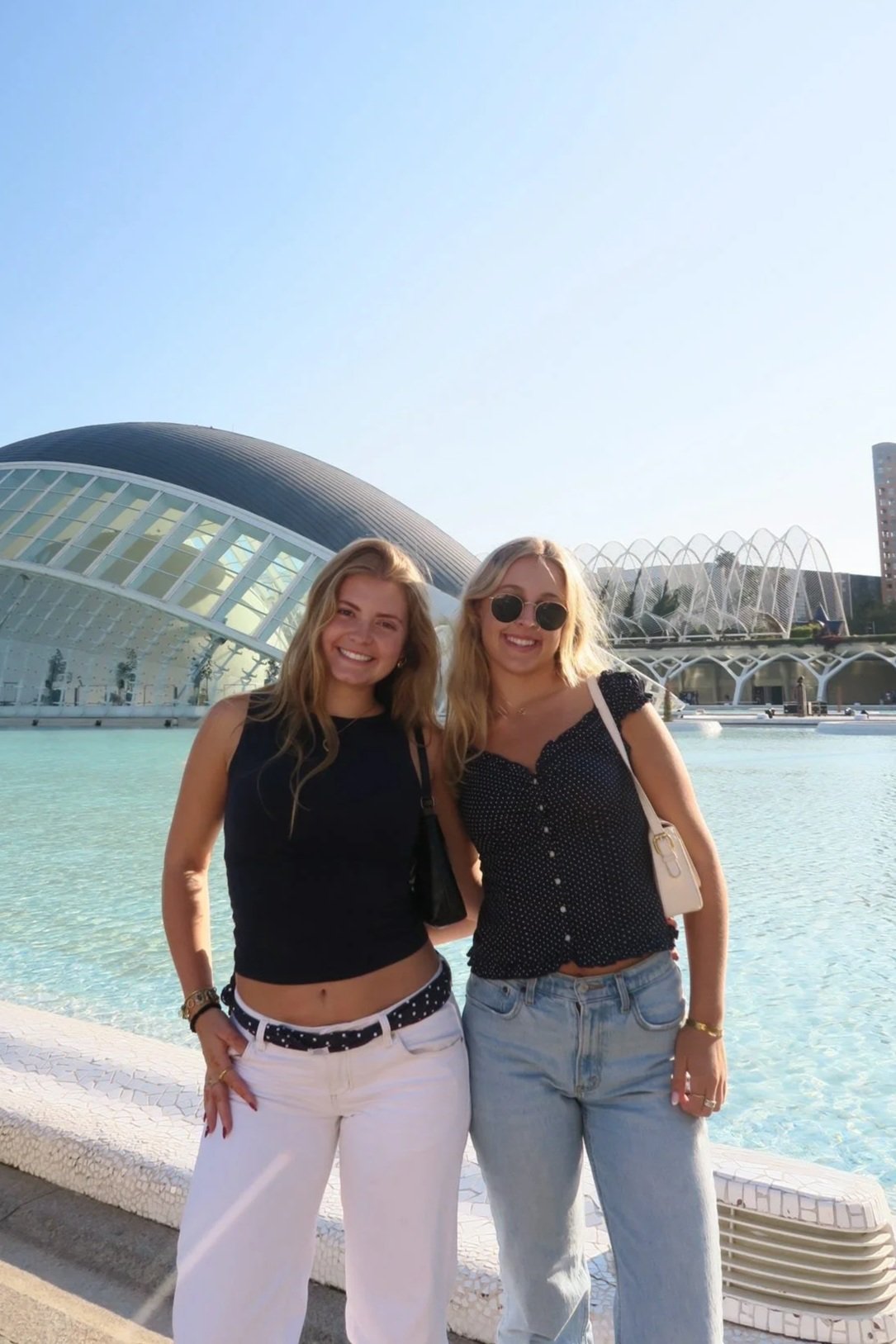 Two young women smiling and standing together in front of the futuristic L'Hemisfèric building and a water feature at the City of Arts and Sciences in Valencia, Spain.