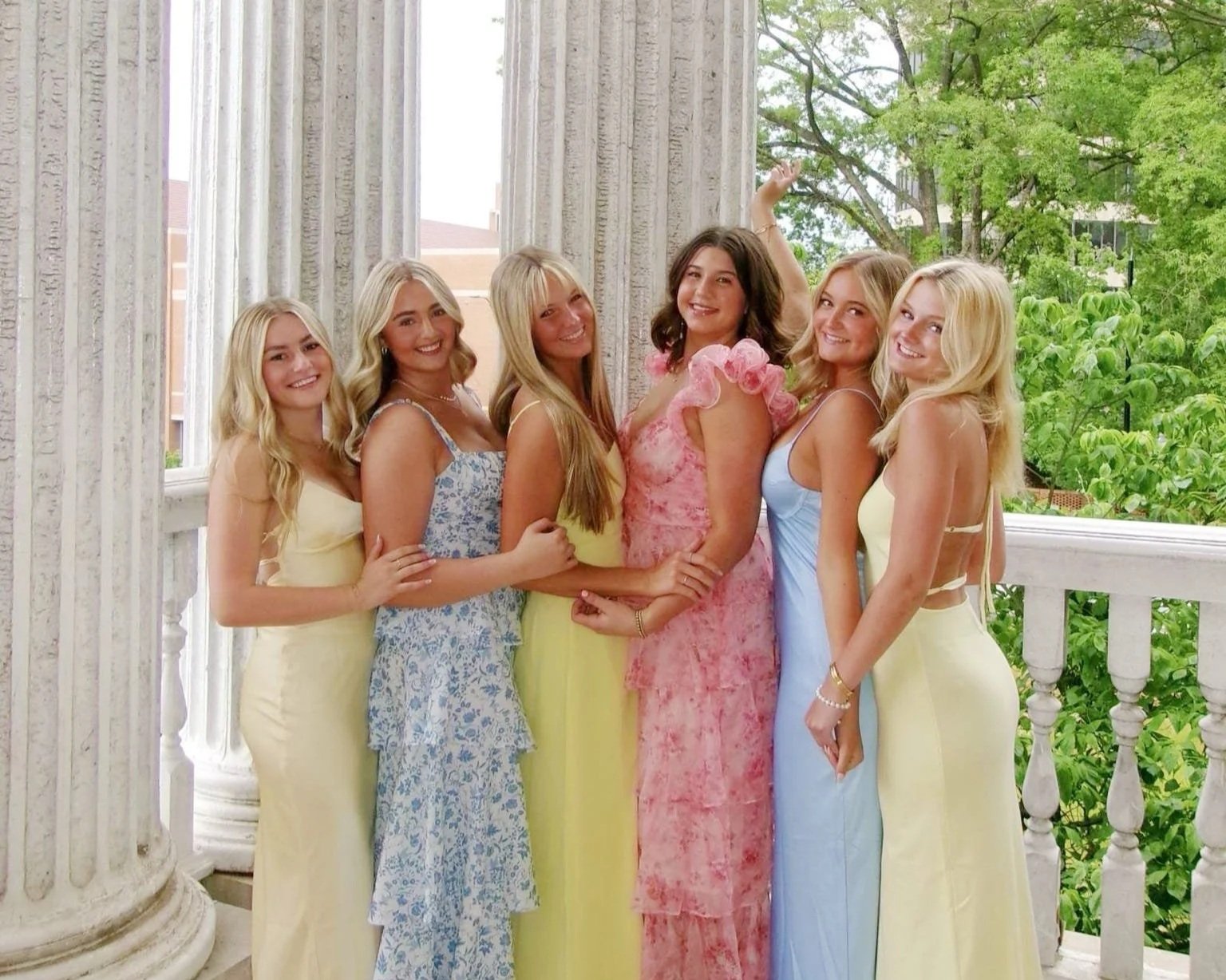 Group of six women dressed in pastel-colored dresses standing on a porch with white columns and green trees in the background.