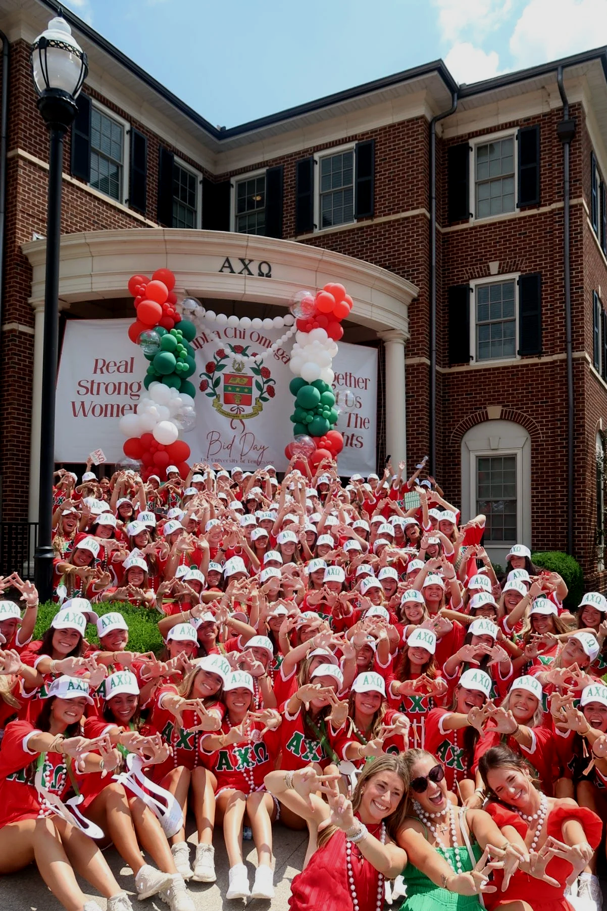 A large group of women dressed in red shirts and white hats with Greek letters, celebrating on the steps of a brick building decorated with red, white, and green balloons, during a fraternity sorority campus event.