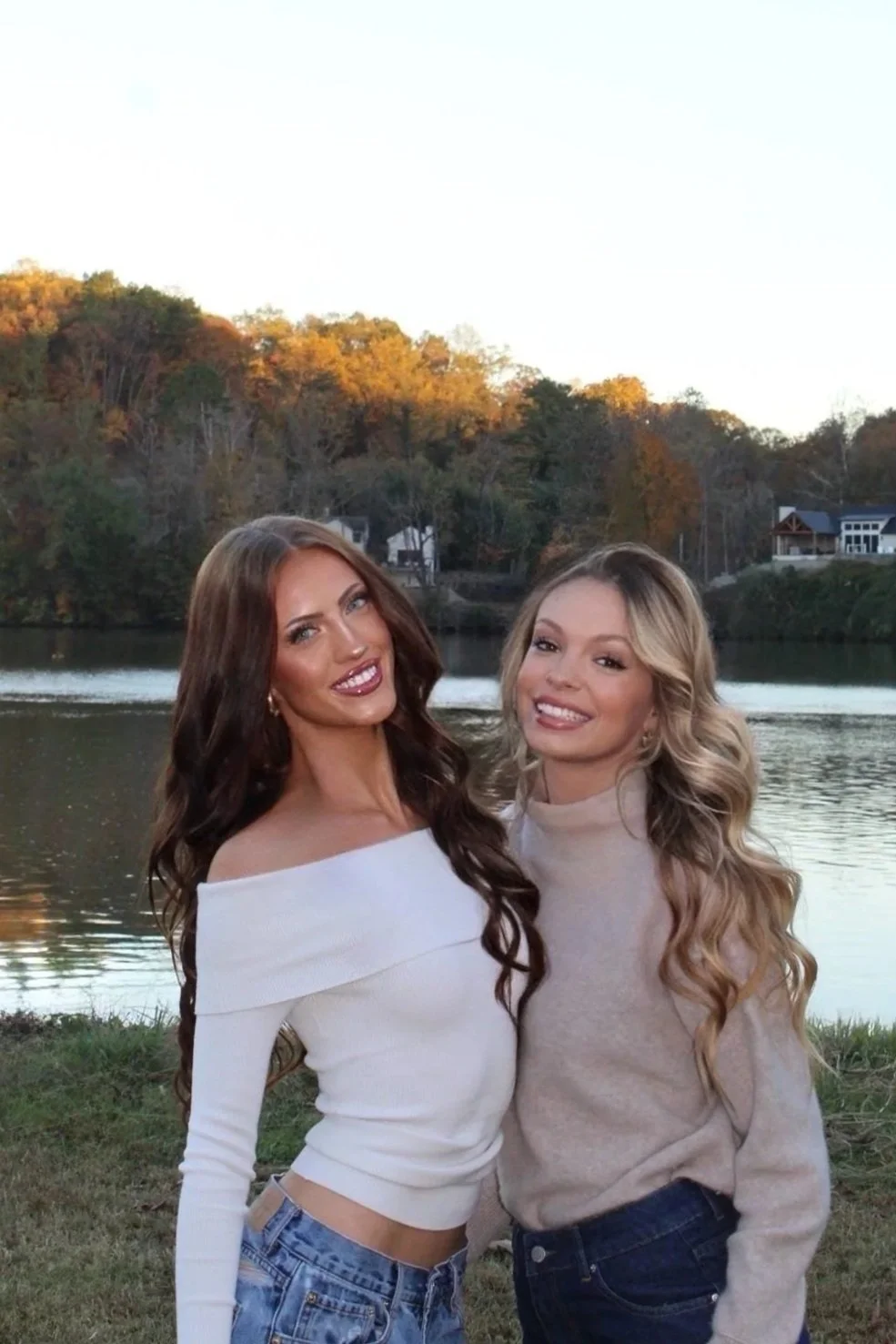 Two young women standing outdoors by a lake with trees and houses in the background, smiling at the camera.