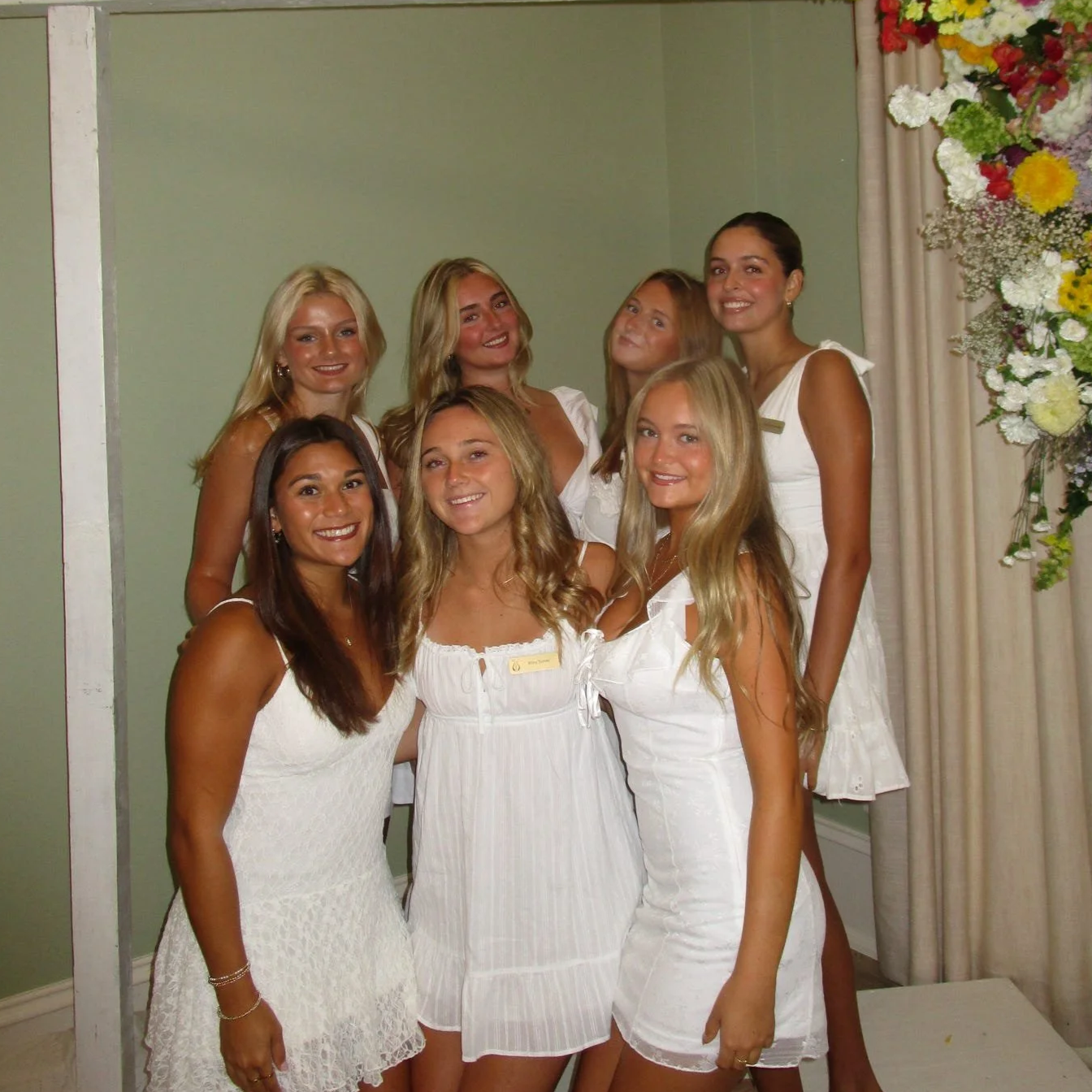 Seven young women dressed in white and smiling for a group photo indoors, with a green wall and floral arrangement in the background.