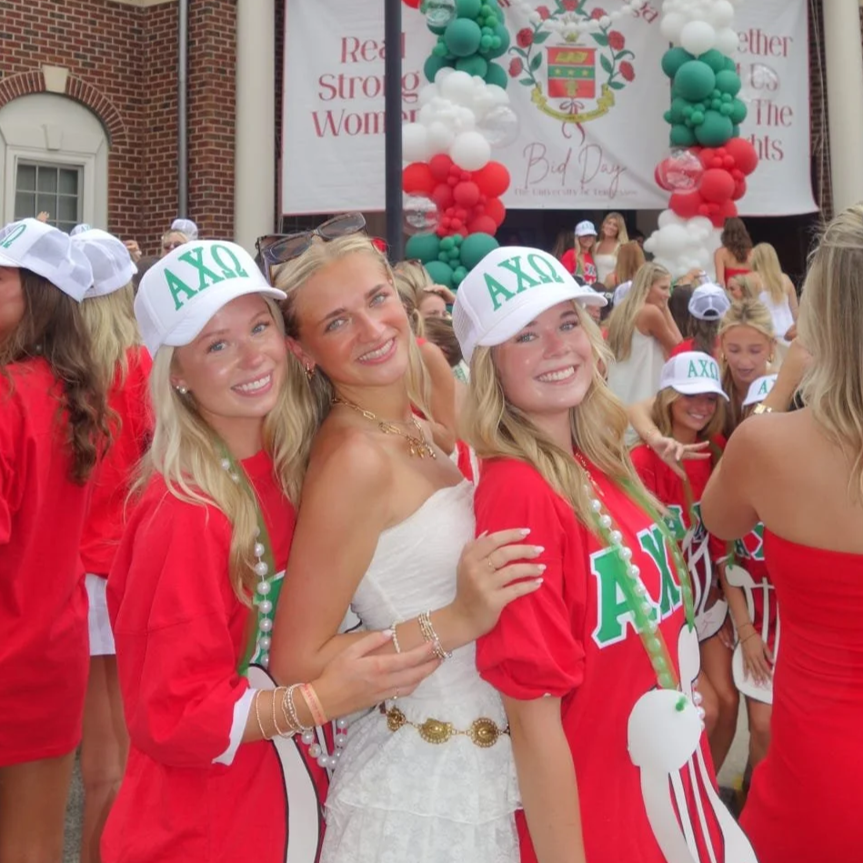 Group of young women celebrating a sorority event, some wearing hats with Greek letters and festive shirts, standing in front of a decorated balloon arch and a banner that reads 'Red, Strong Women, Bad Day'.