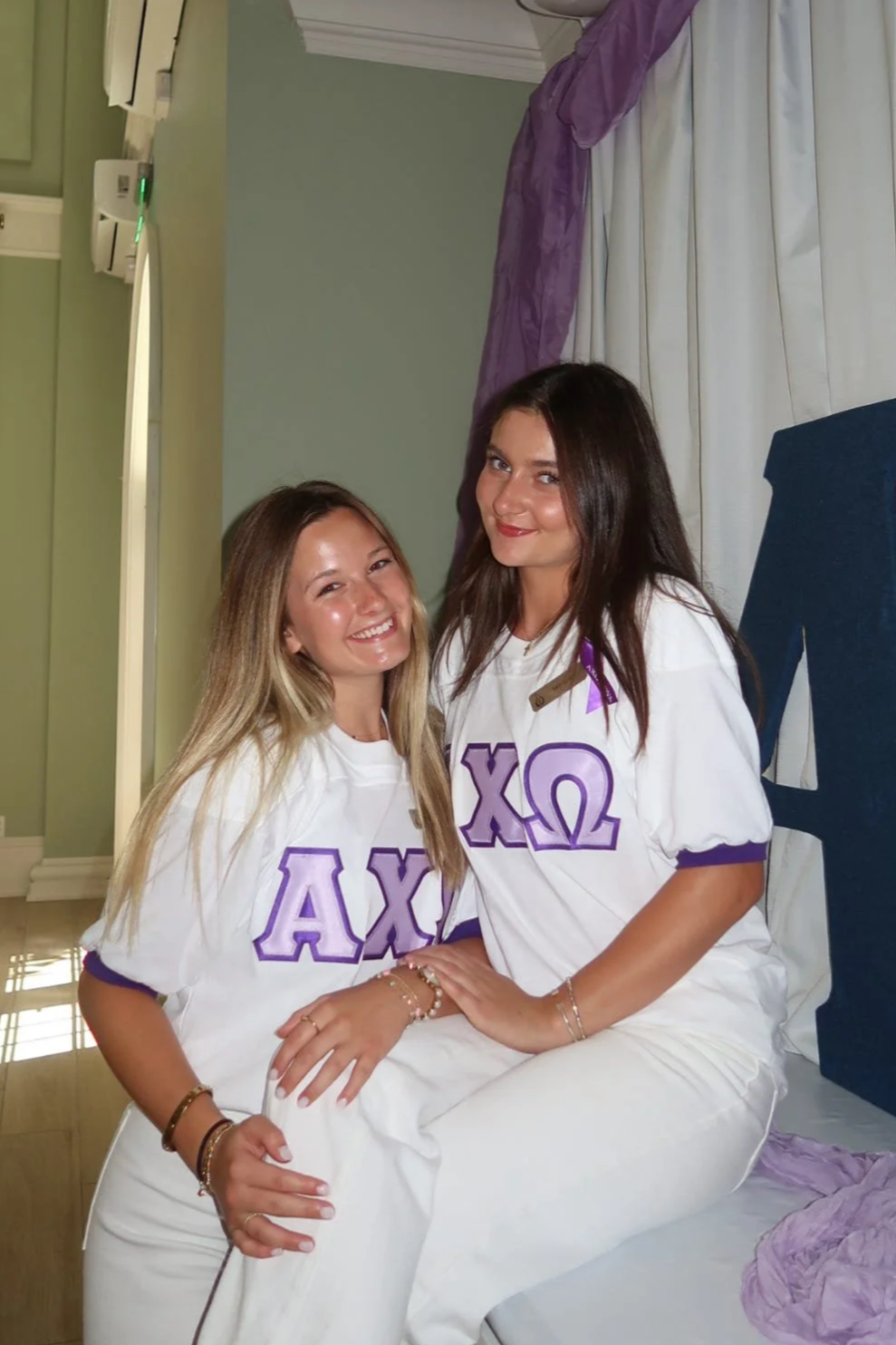 Two young women sitting on a bed, smiling and wearing sorority shirts with Greek letters in purple and white.