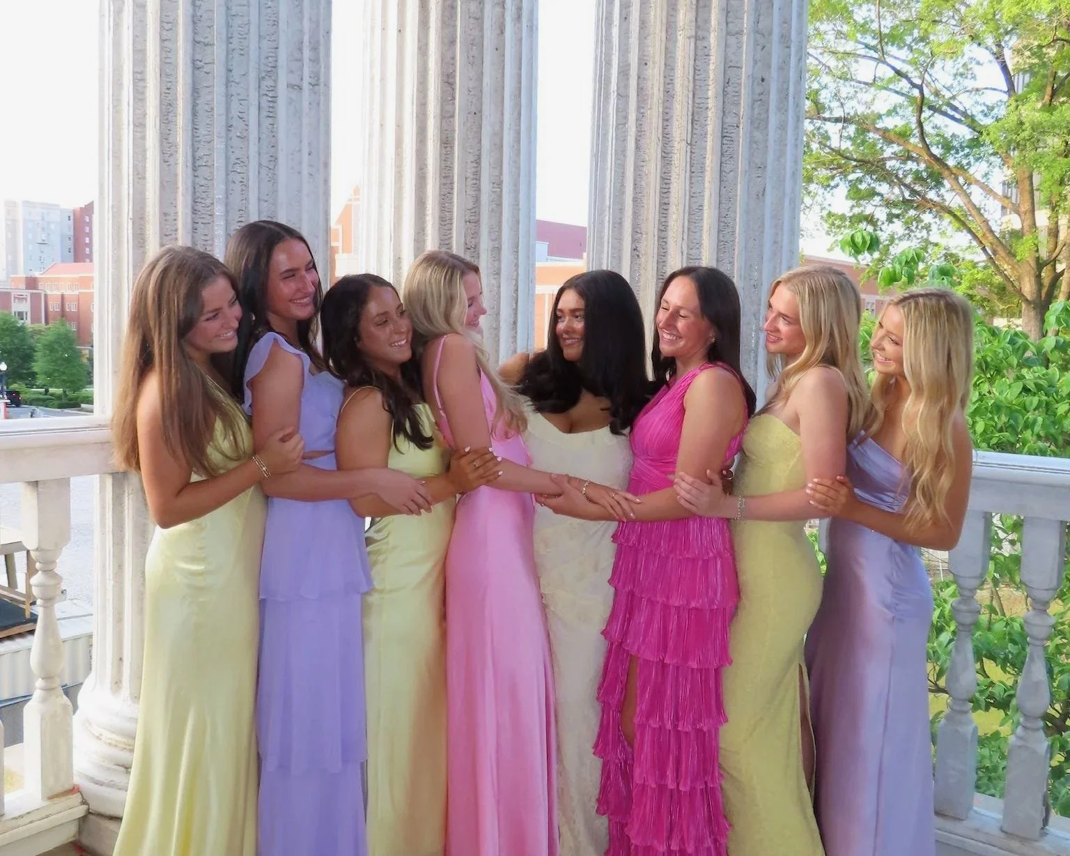 Group of women dressed in colorful dresses standing together and smiling on a balcony with columns and a view of trees and buildings outside.
