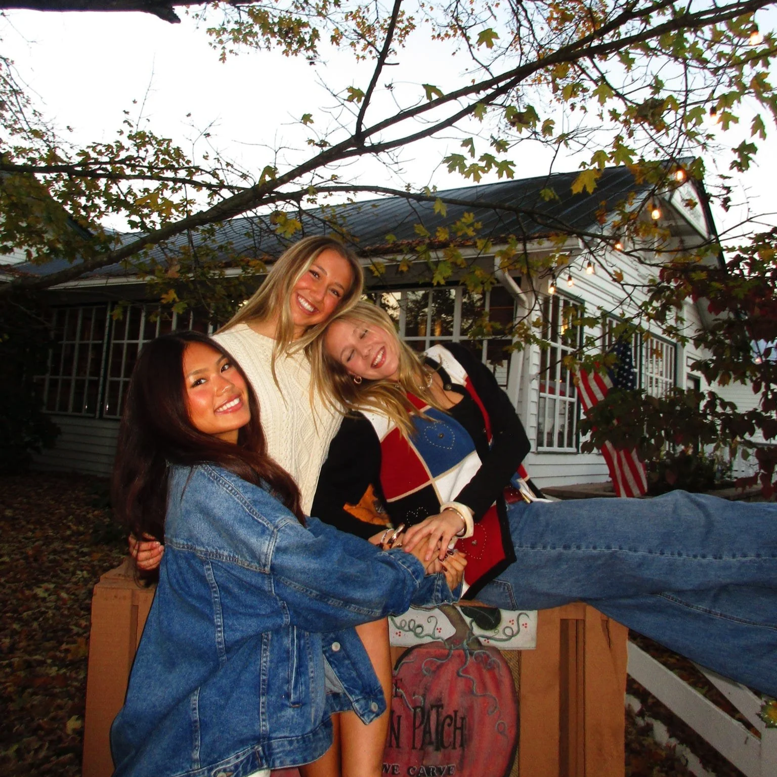 Three young women smiling and holding hands outdoors in front of a house decorated for fall, with autumn leaves on the ground and string lights overhead.