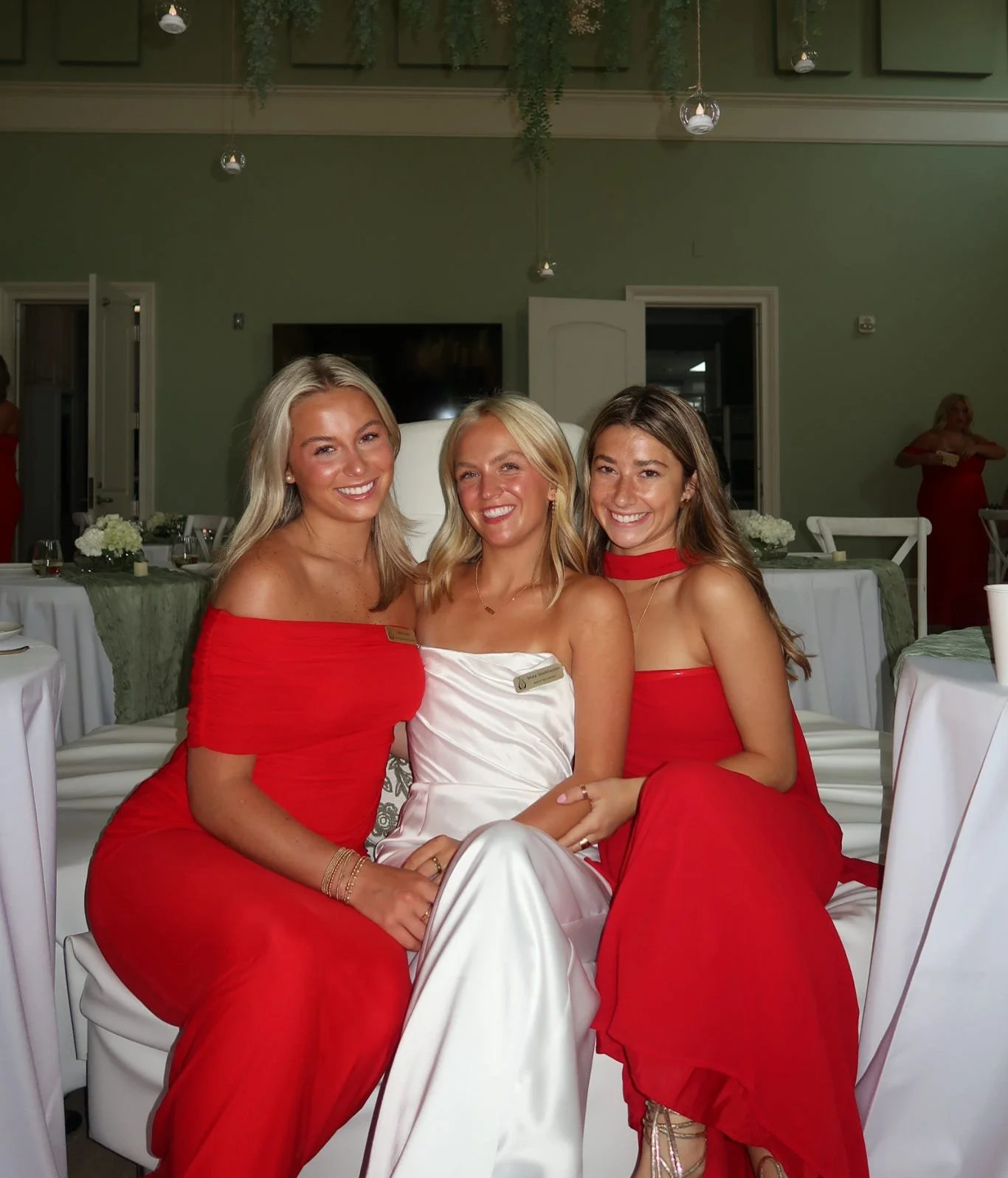 Three women sitting together at a formal event in a decorated room. They are smiling and wearing elegant outfits, with the women on the left and right in red dresses and the woman in the middle in a white dress.