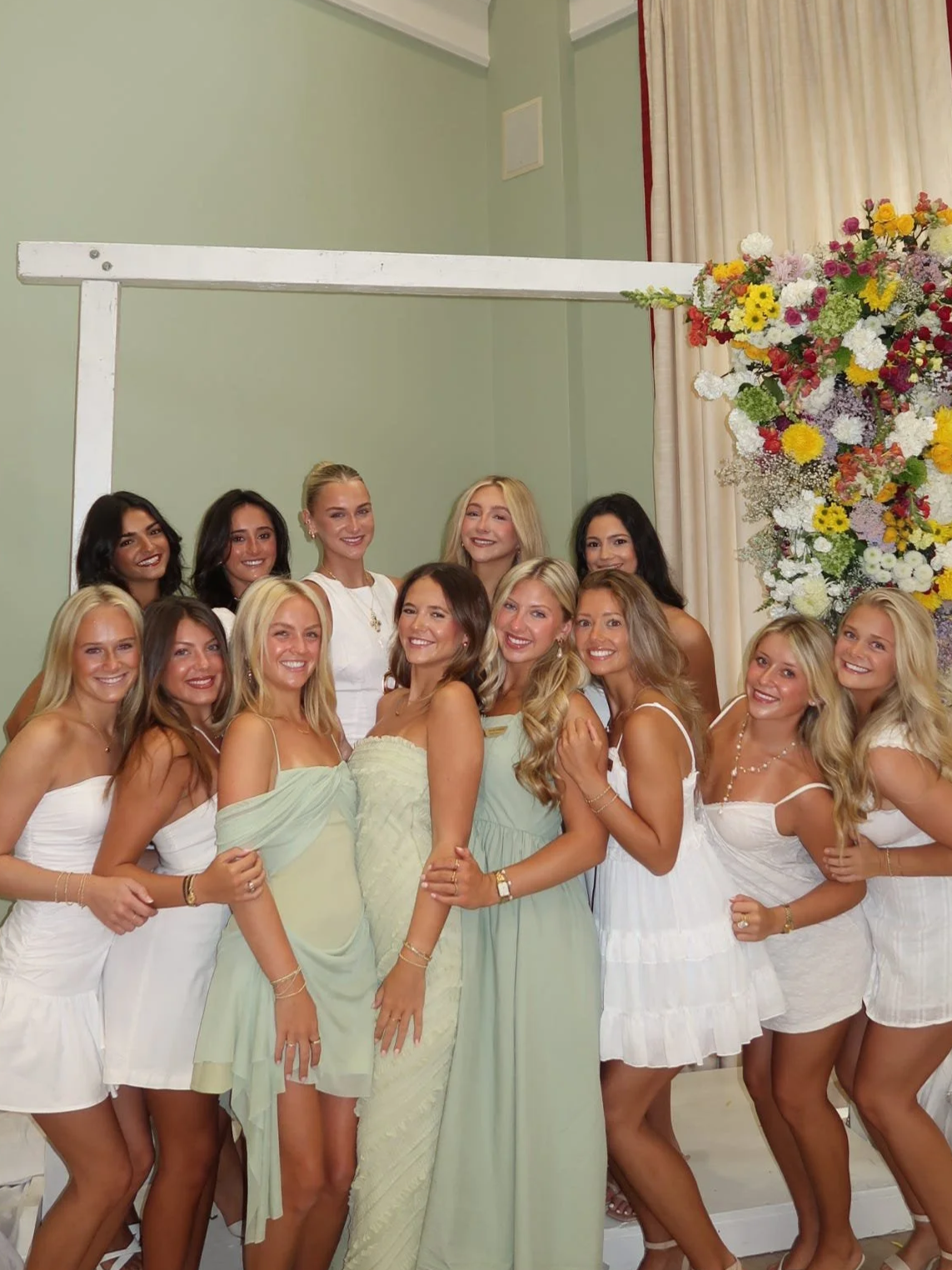 A group of women dressed in light-colored dresses, standing together and smiling in front of a large floral arrangement with white, yellow, pink, and purple flowers, at a celebration or event.