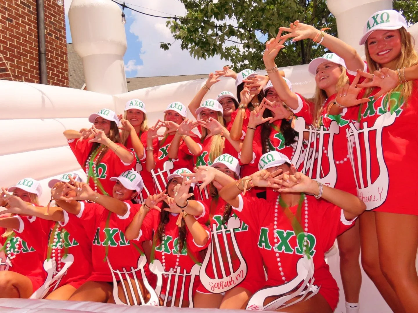 Group of young women dressed in red shirts with sorority symbols, wearing white hats with Greek letters, celebrating and making heart signs with their hands at an outdoor event.