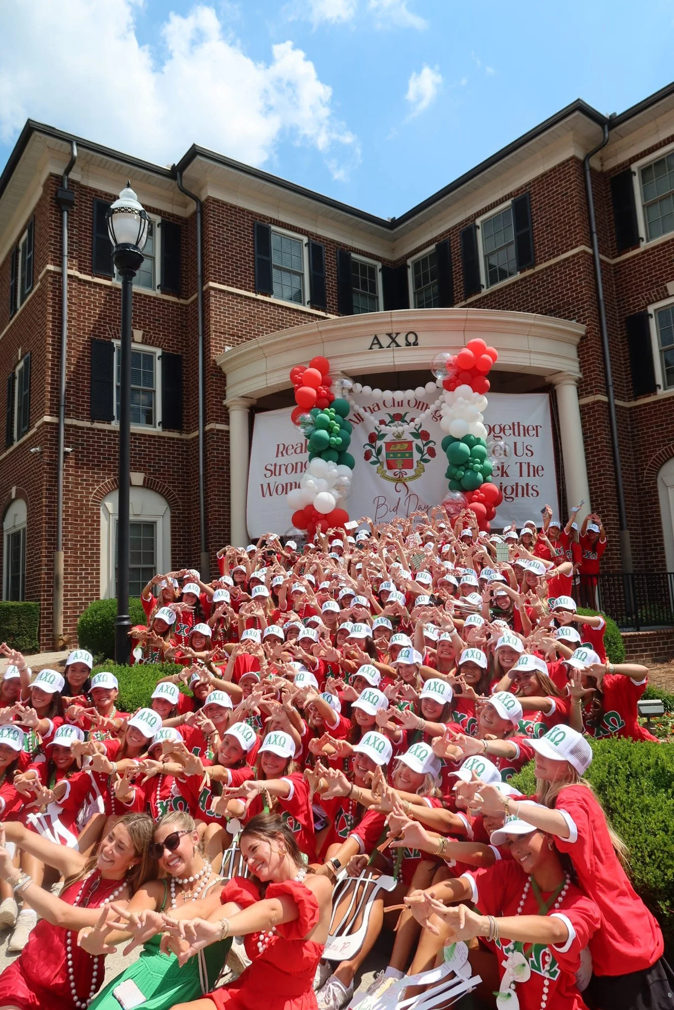 Large group of students dressed in red shirts and white hats with Greek letters, posing on the steps in front of a large brick building with Greek letters, decorated with red, white, and green balloons, celebrating a sorority event.