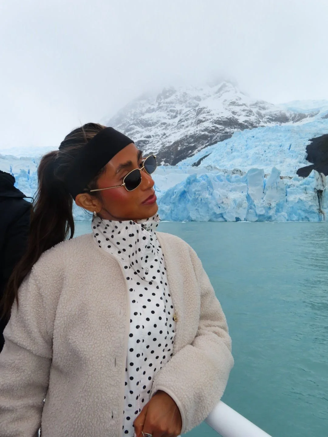 Woman wearing a cream fleece jacket, polka dot scarf, and sunglasses at a glacier with snowy mountains in the background.