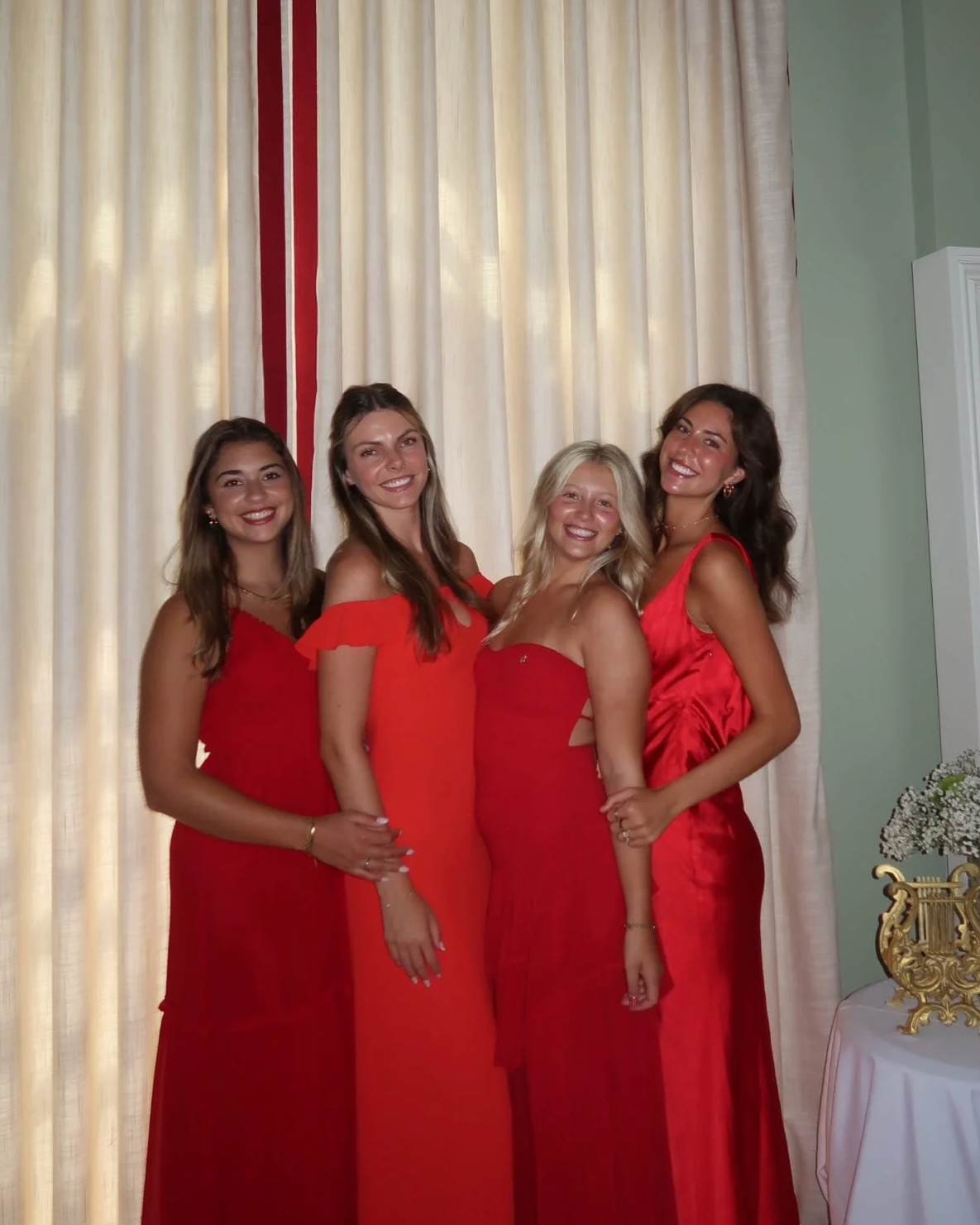 Four women in red dresses standing together and smiling in front of cream-colored curtains.